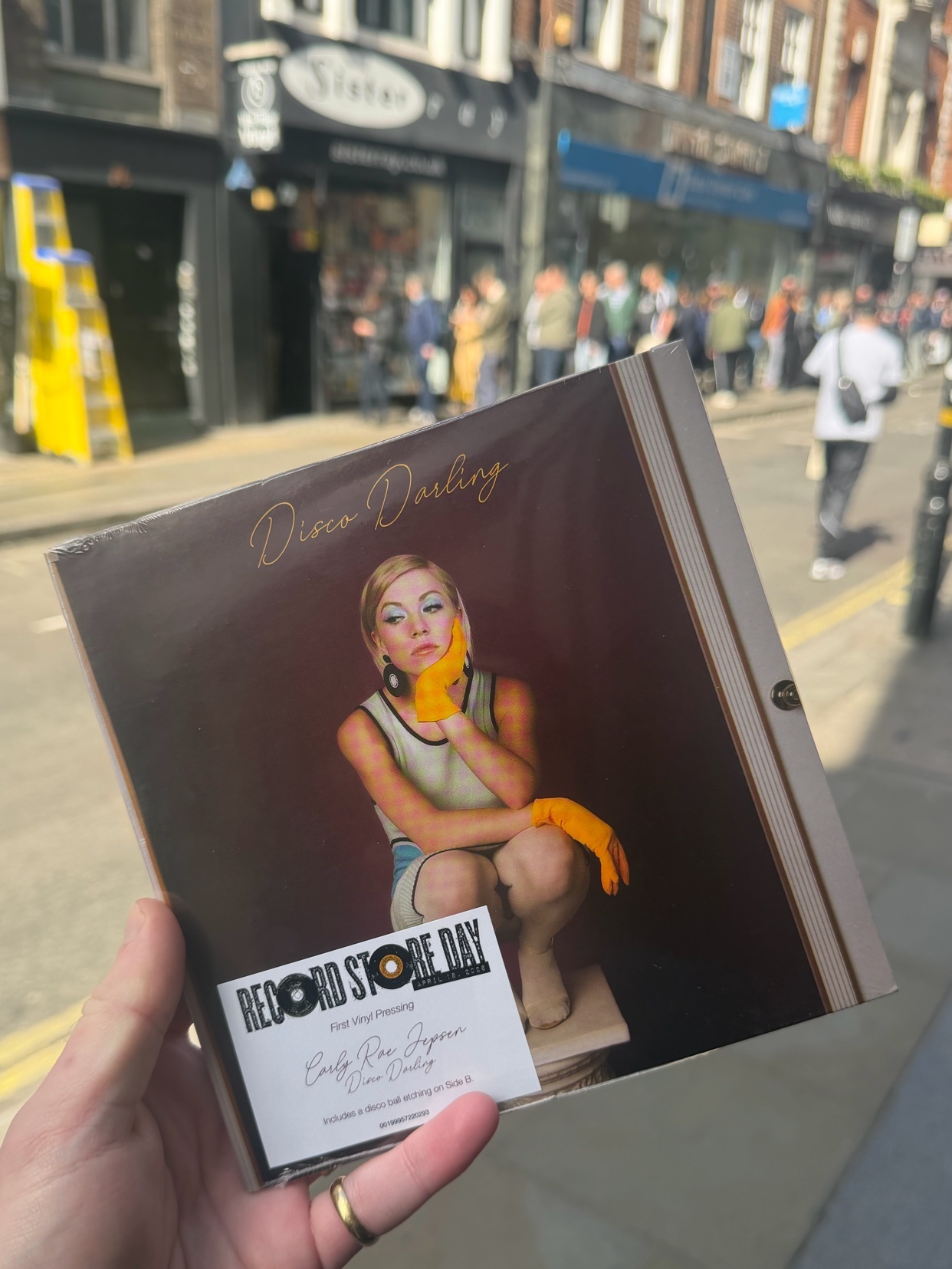 A hand holds a vinyl record sleeve featuring a woman in yellow gloves and a vintage-inspired outfit. The background shows a bustling street scene, likely in Soho, London, during Record Store Day.