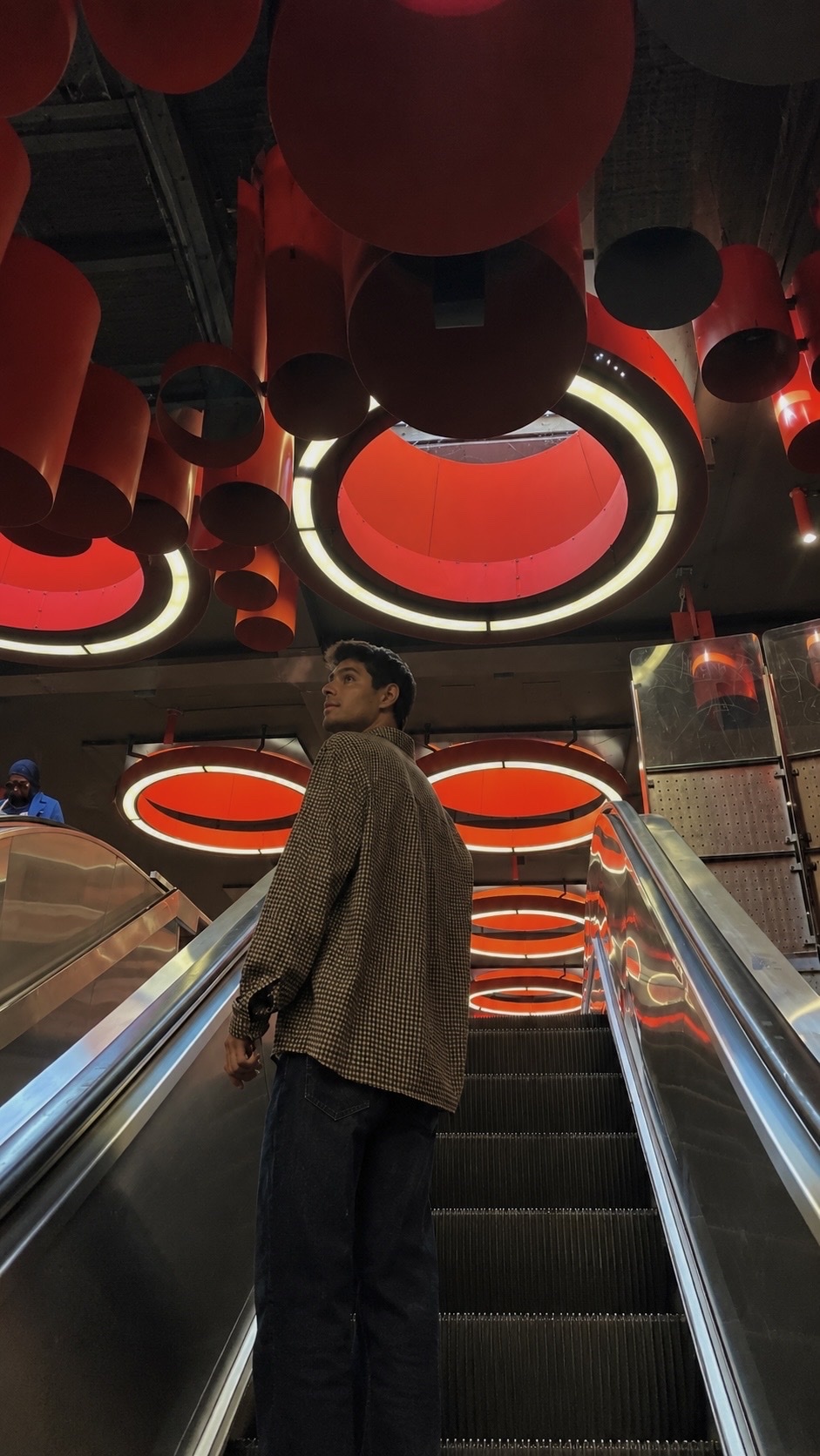 A man rides an escalator in a modern subway station with striking red cylindrical light fixtures overhead.