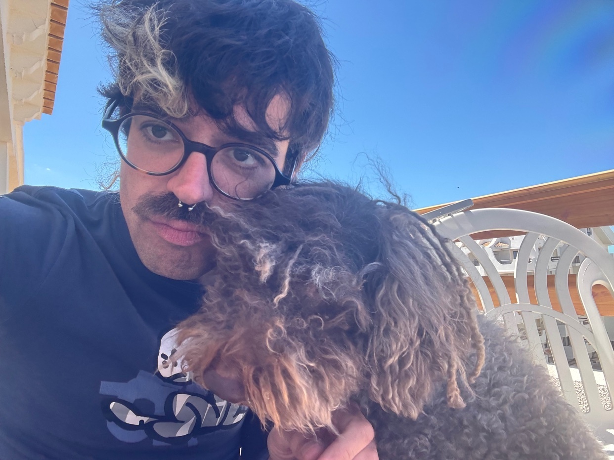 A man with a mustache and glasses poses for a selfie with his fluffy brown dog. The man has dark curly hair with a blonde streak and a nose ring. The dog has curly, matted fur and rests its head on the man's shoulder.