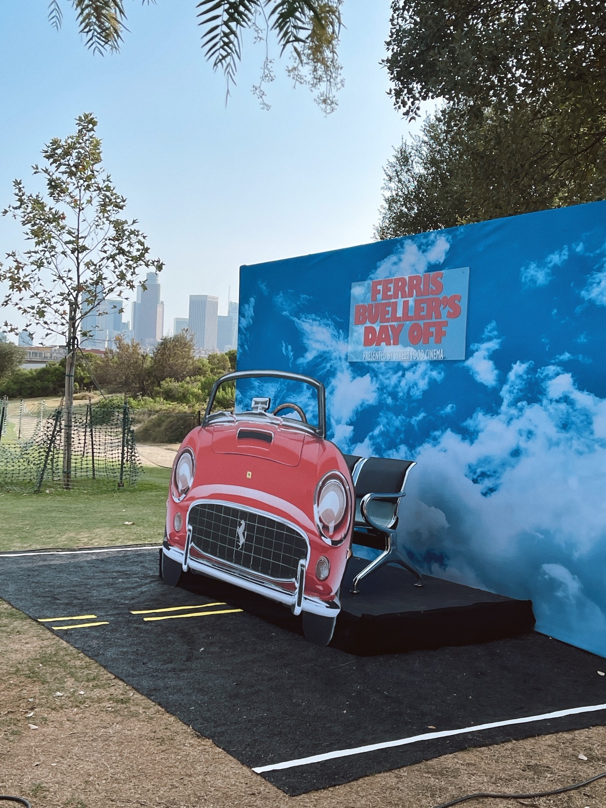 A cardboard cutout of a red Ferrari convertible is displayed outdoors with a backdrop featuring clouds and the title 'Ferris Bueller's Day Off'. The display is set up in a park-like area with trees and a cityscape visible in the distance.
