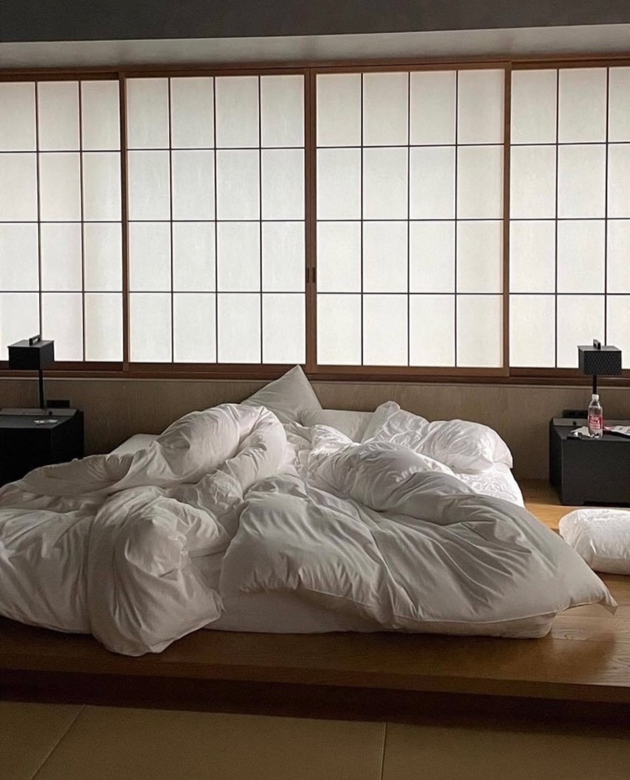 A minimalist bedroom with a rumpled white bed and a traditional Japanese-style window. The room has a serene and uncluttered aesthetic.