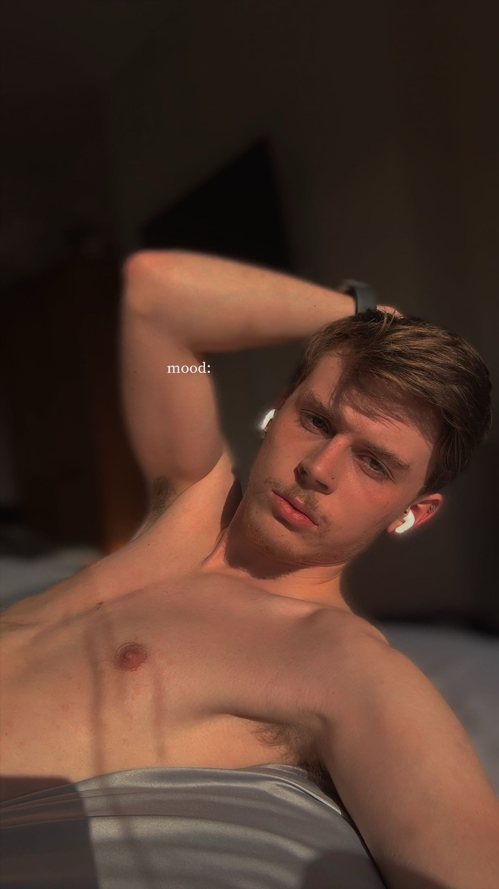 A shirtless man with stubble and a mustache lies on a bed, bathed in warm sunlight. He is wearing earbuds and has one arm behind his head.
