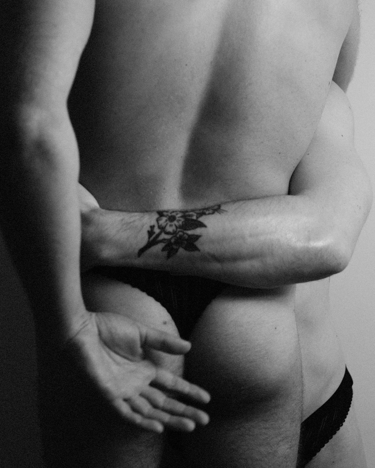 A black and white photo of two men embracing. One man is shirtless, and the other has his arm around him.