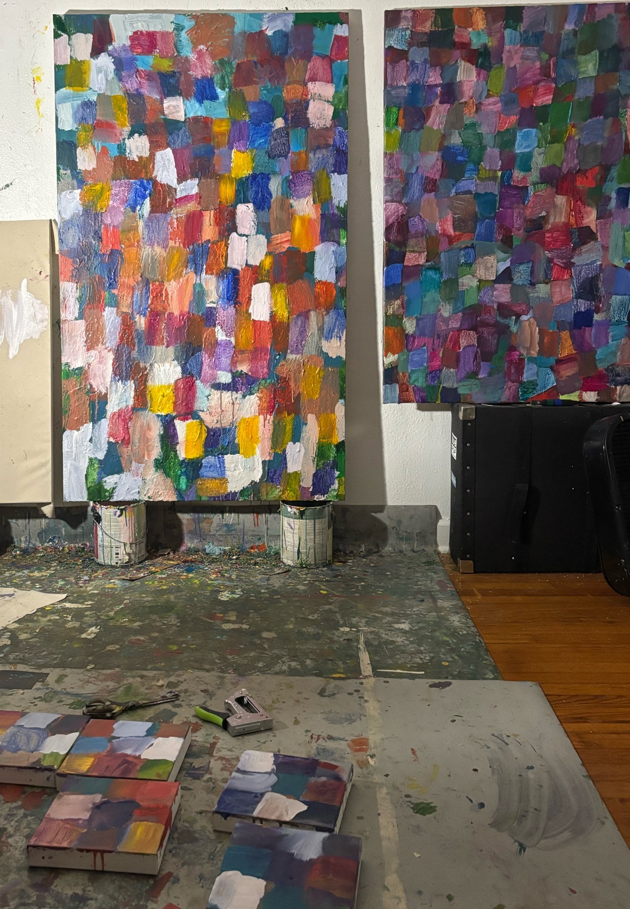 Two large, colorful abstract paintings hang on a wall in an art studio. Smaller canvases with similar abstract patterns are laid out on a table in the foreground.