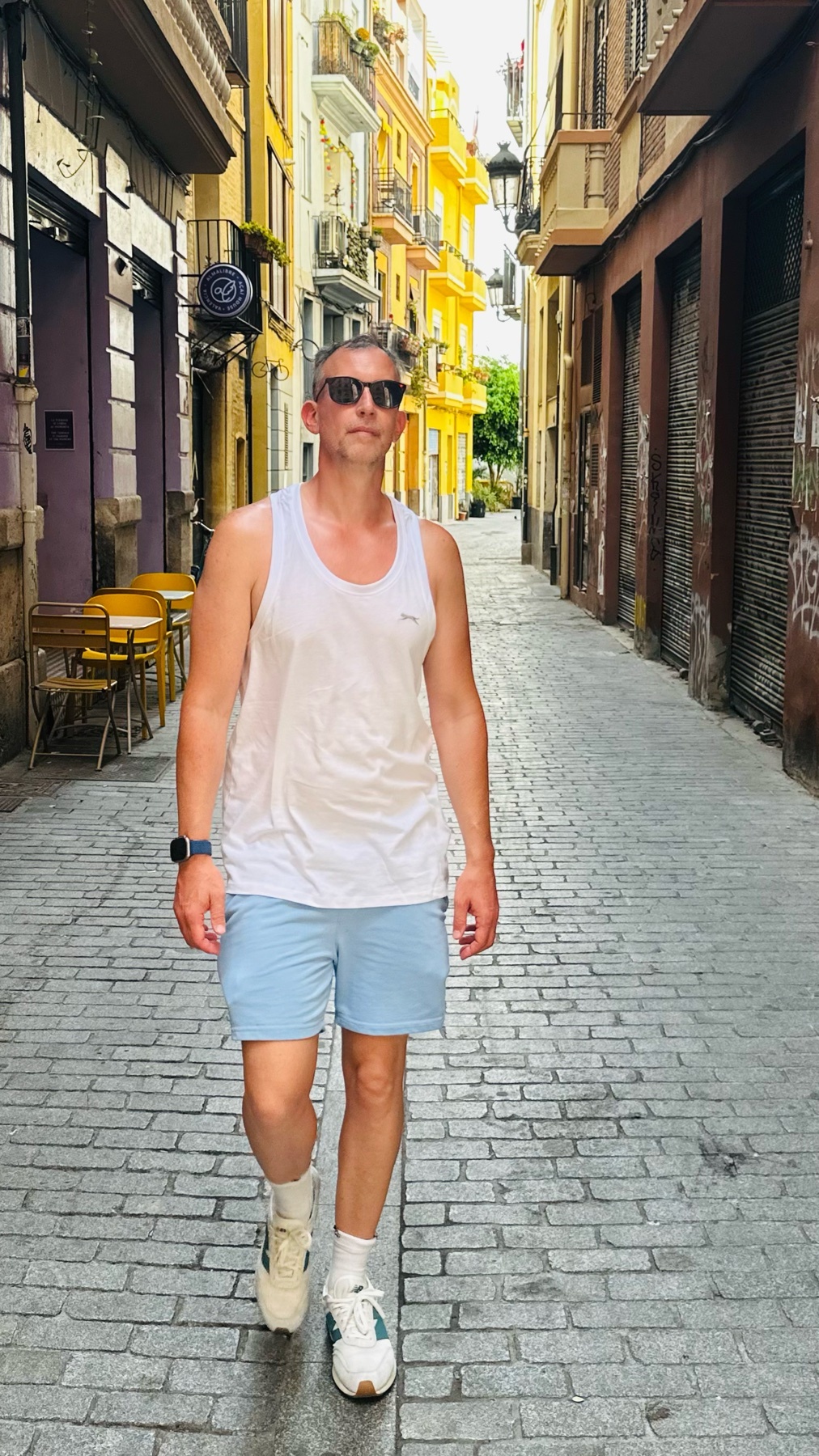A man walks down a narrow, cobblestone street in Valencia, Spain. He is wearing a white tank top, light blue shorts, sunglasses, and sneakers.