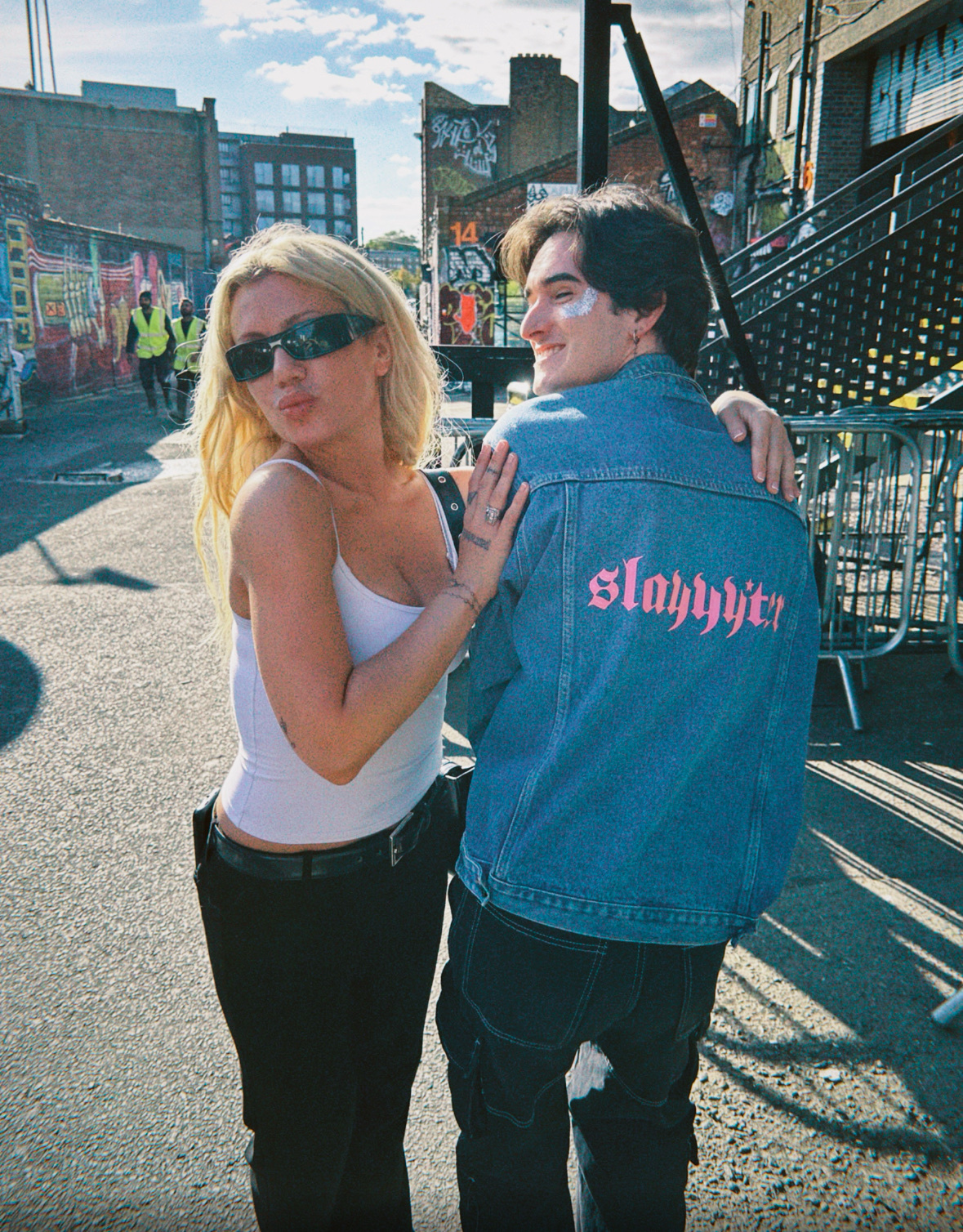 A blonde woman wearing sunglasses and a white tank top poses with a man in a denim jacket. They are standing outdoors with a city backdrop.