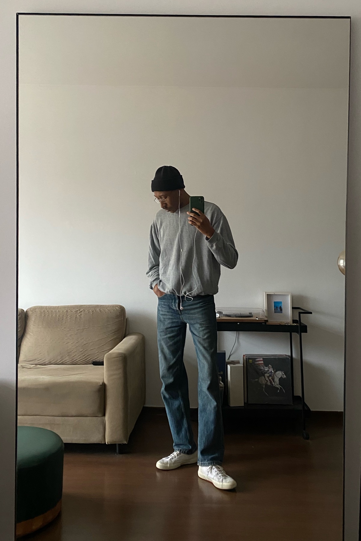 A man takes a mirror selfie in a casual outfit, wearing a beanie, sweatshirt, jeans, and sneakers.