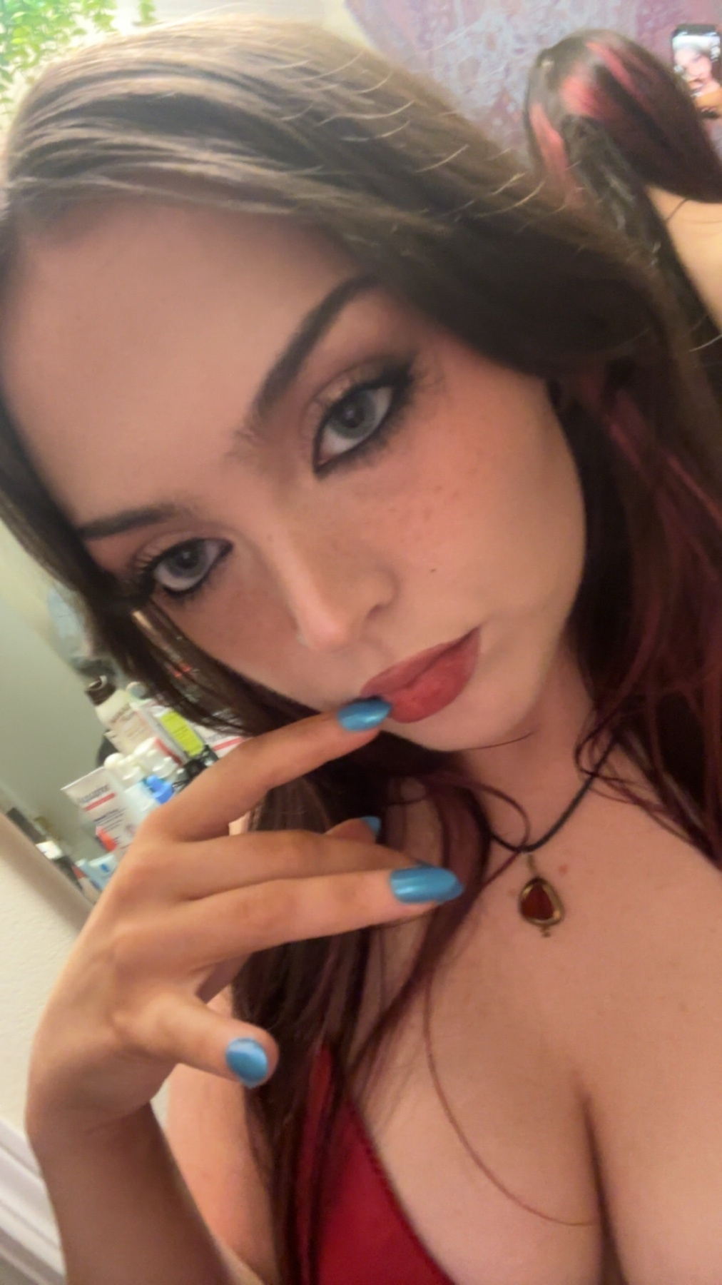 A woman takes a selfie, showcasing her makeup and red hair. She is wearing a necklace and has blue nail polish.