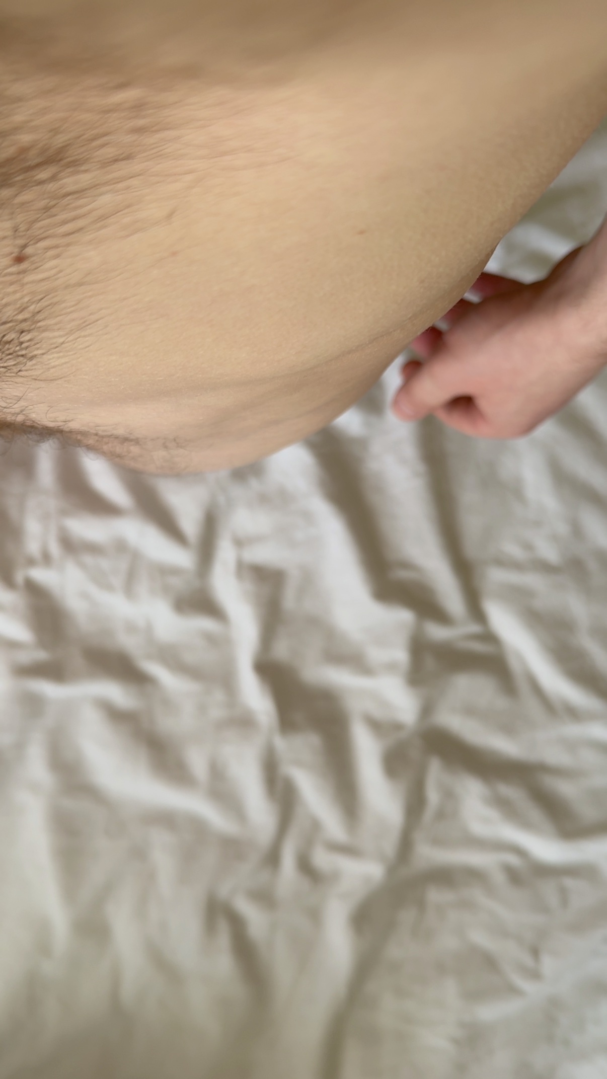 A close-up shot of a person's lower body, focusing on body hair and skin against white, wrinkled bed sheets. A hand is visible reaching towards the body hair.
