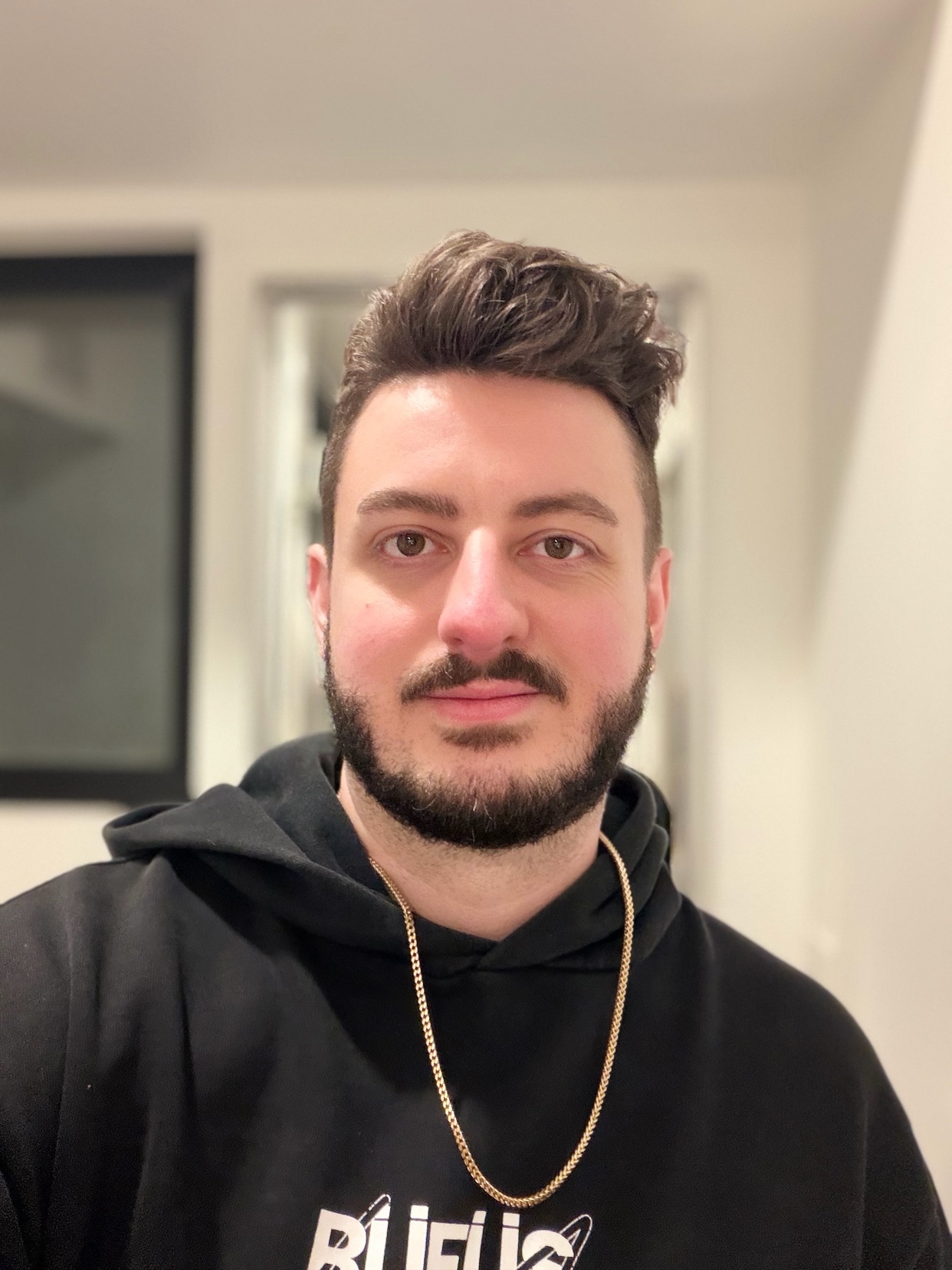 A man with dark hair and a beard wears a black hoodie and a gold chain.