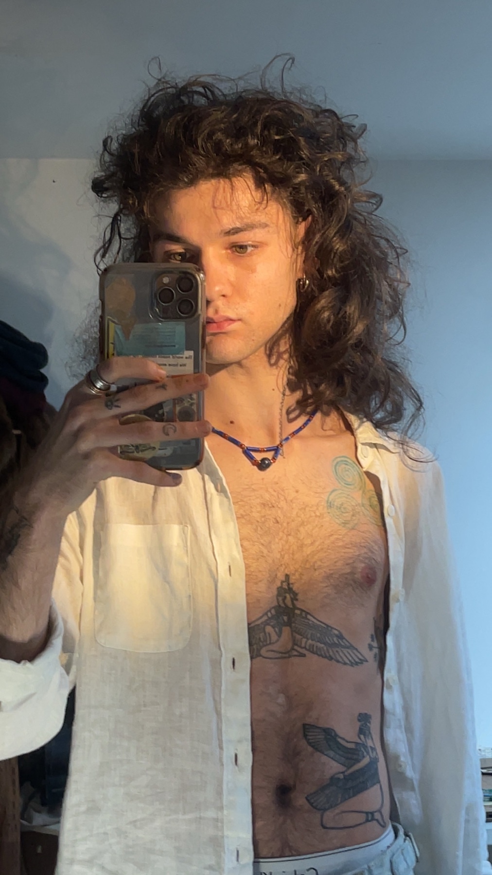 A man takes a mirror selfie, shirtless and wearing an open white shirt. His chest and torso are covered in tattoos.