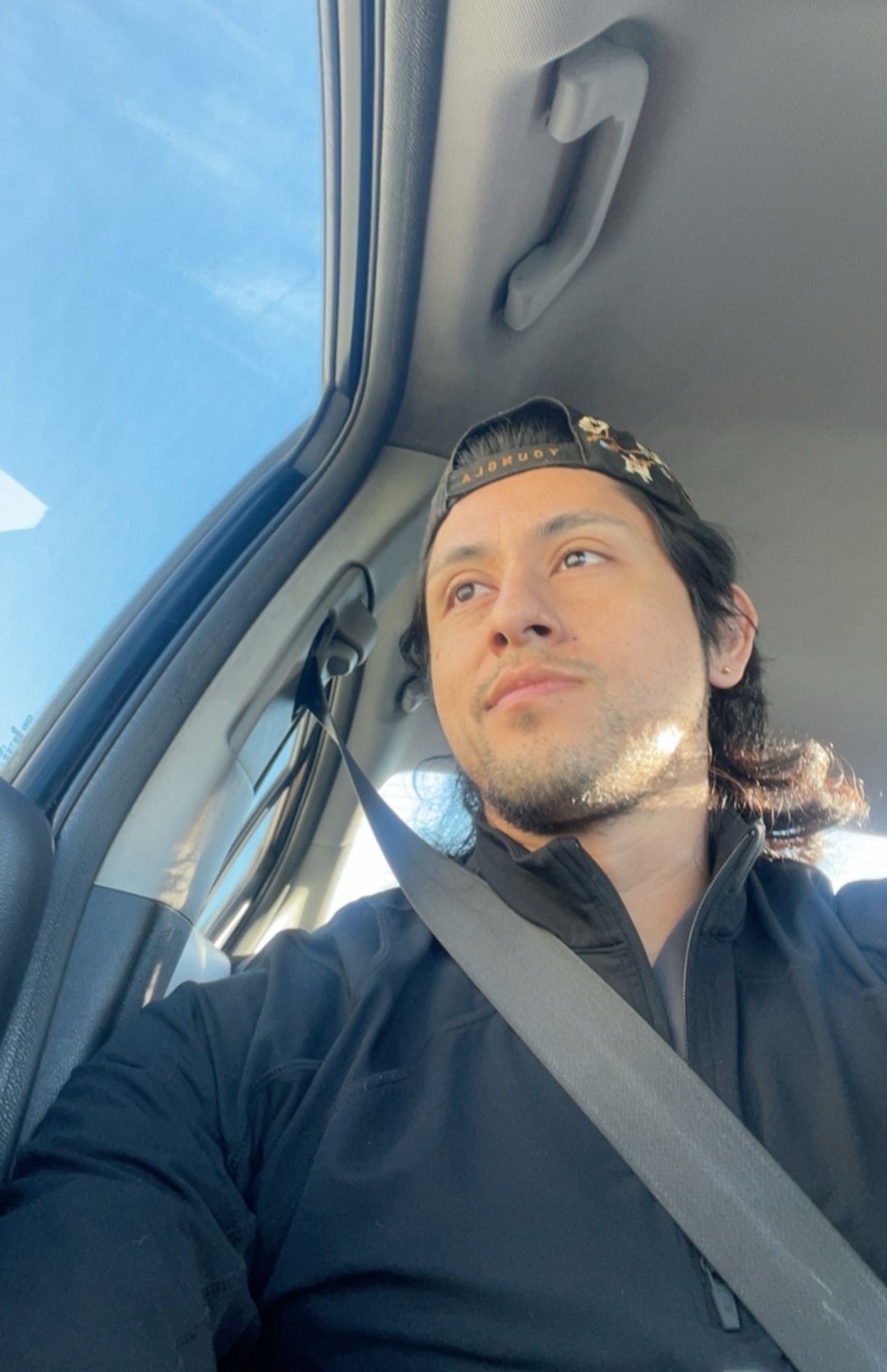 A man wearing a black baseball cap and a black jacket is captured in a selfie taken from inside a car. The sun is shining brightly, casting a warm glow on his face as he looks off to the side.