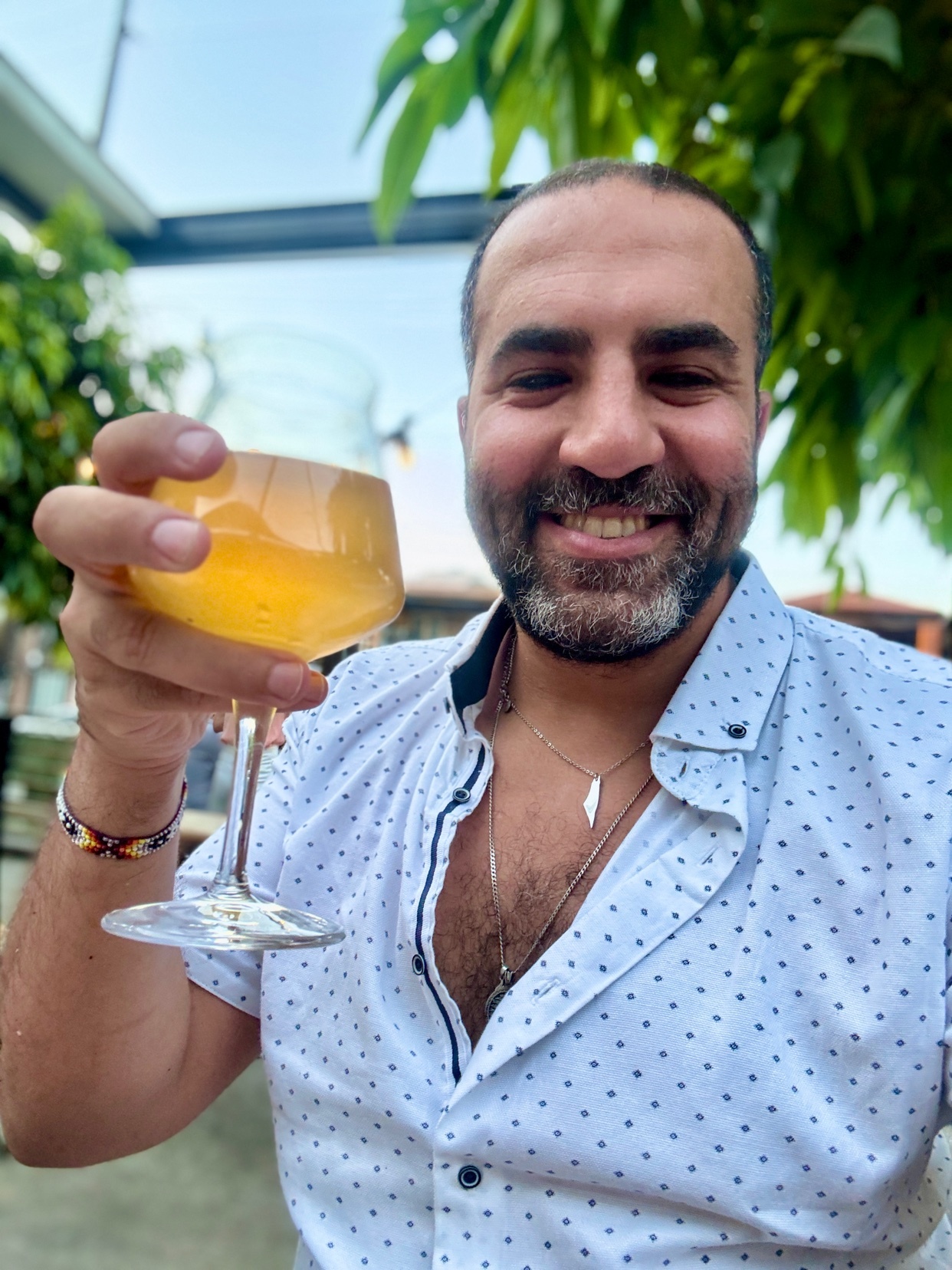 A man with a beard and stubble smiles as he holds up a glass of yellow liquid. He is wearing a white short-sleeved shirt with blue polka dots and a necklace.