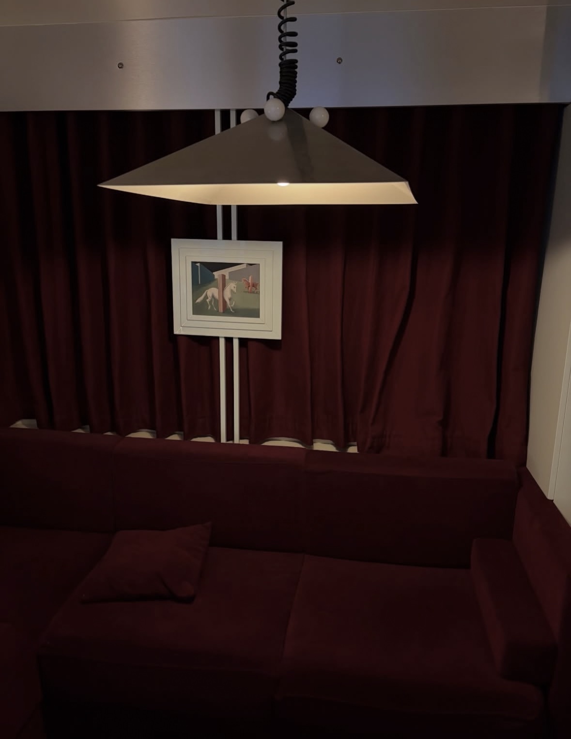 A modern living room features a deep red sofa and matching curtains, with a unique geometric pendant light hanging overhead. A framed piece of art is displayed against the backdrop of the curtains.