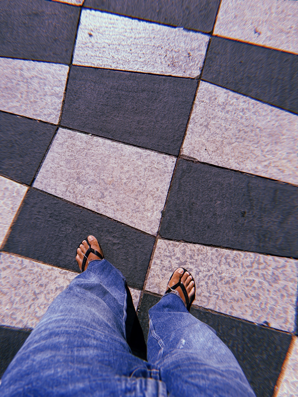 A first-person view of a person wearing blue jeans and flip-flops standing on a black and white tiled floor.