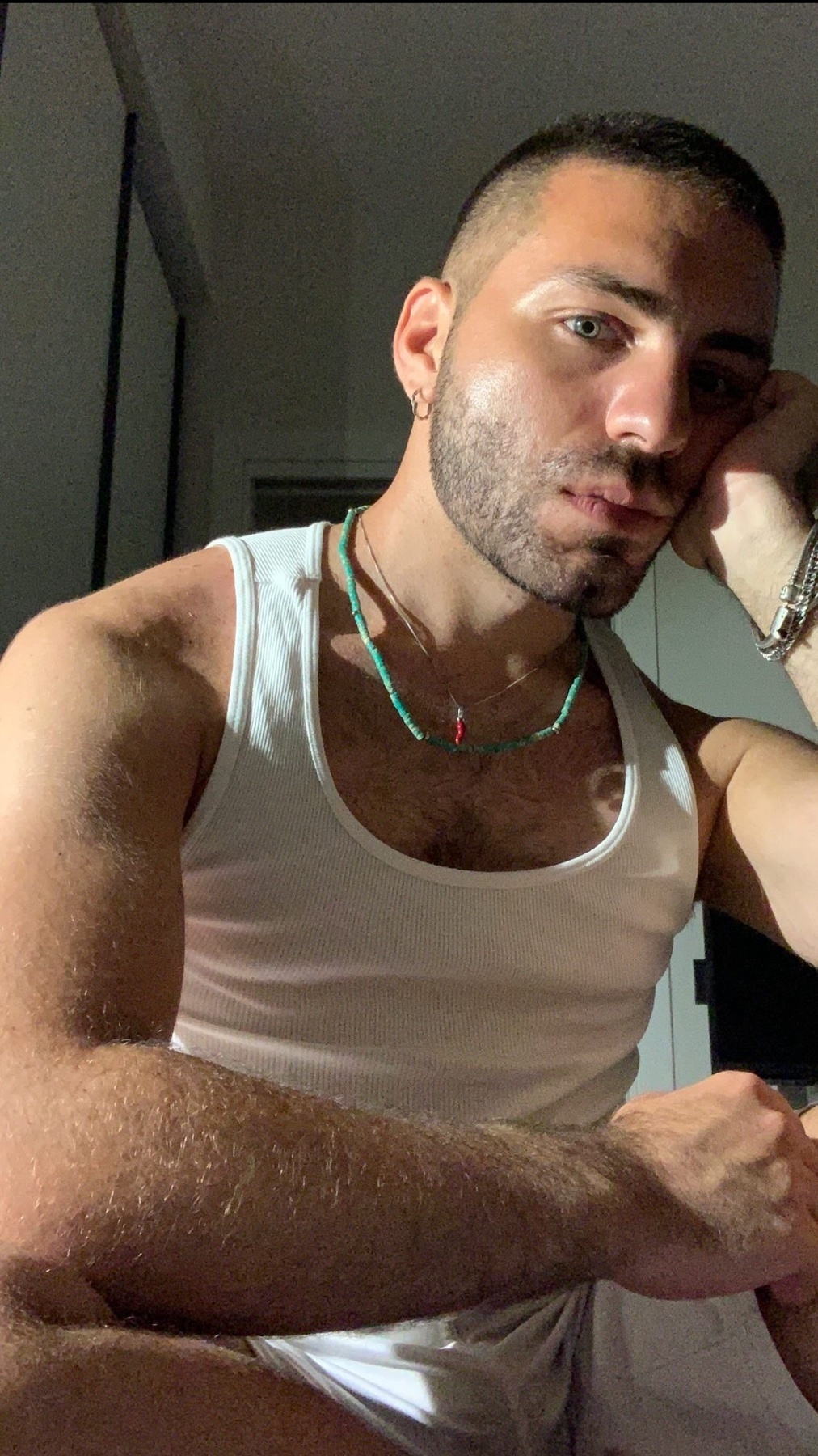 A close-up, low-angle shot of a man with short hair and stubble, wearing a white tank top and a beaded necklace.