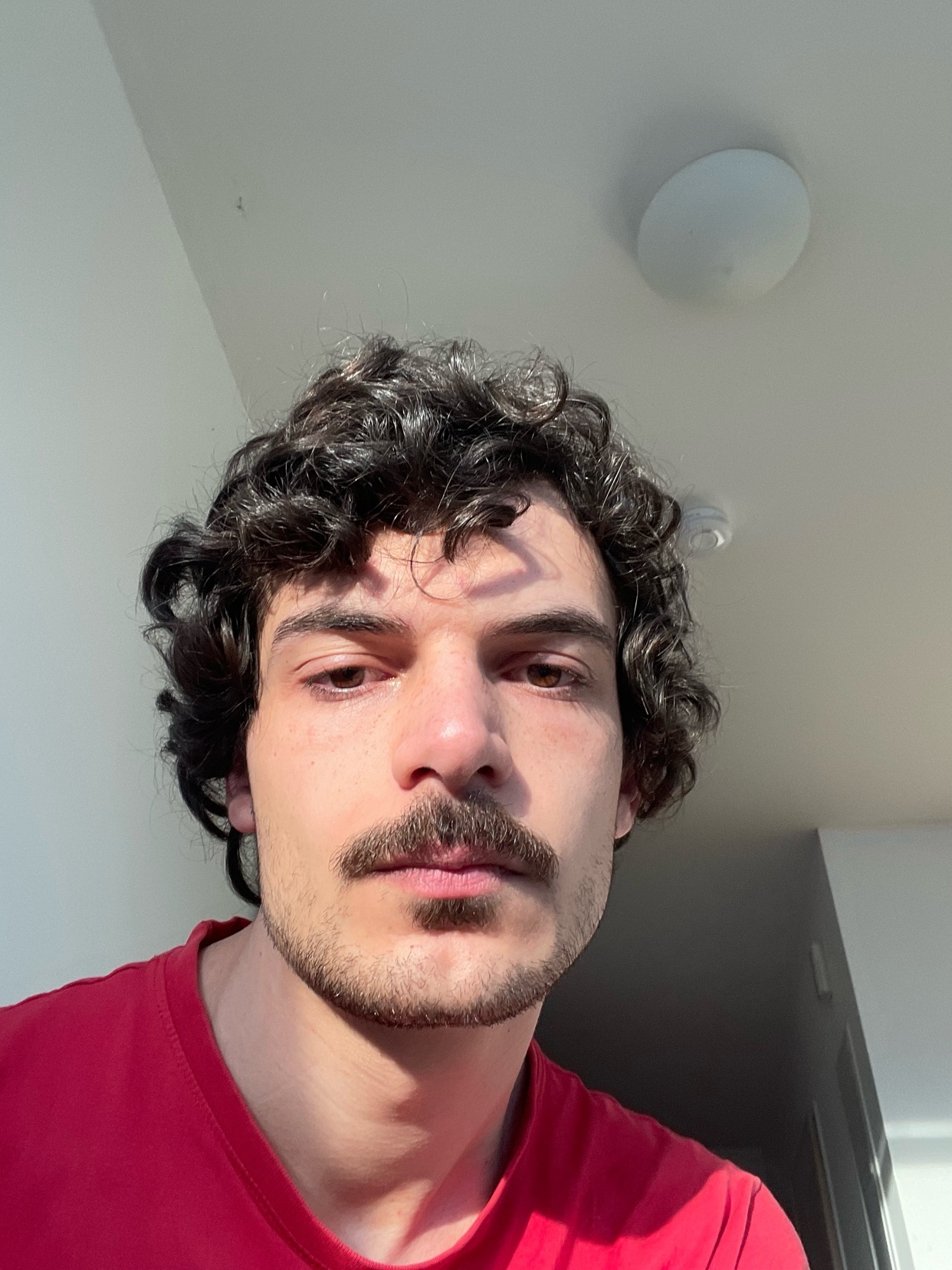 A selfie of a man with curly hair and a mustache. He is wearing a red t-shirt.