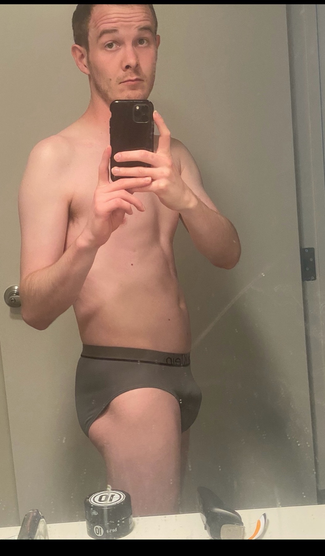 A man in grey briefs takes a mirror selfie in a bathroom.