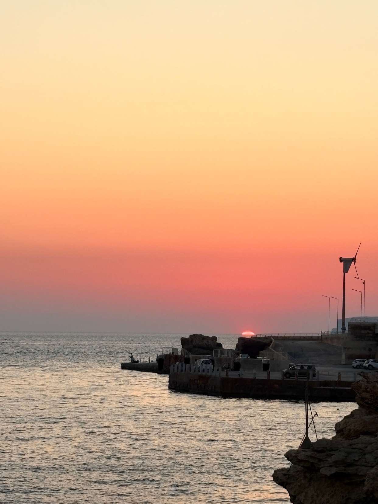 A beautiful sunset paints the sky in hues of orange and pink over a calm ocean. The sun is setting behind a rocky coastline with a pier and a wind turbine visible.