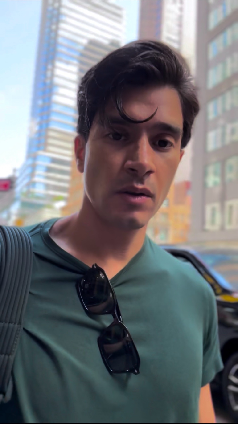 A man with dark hair and sunglasses hanging from his shirt looks into the camera with a serious expression. He is standing outside with buildings in the background.