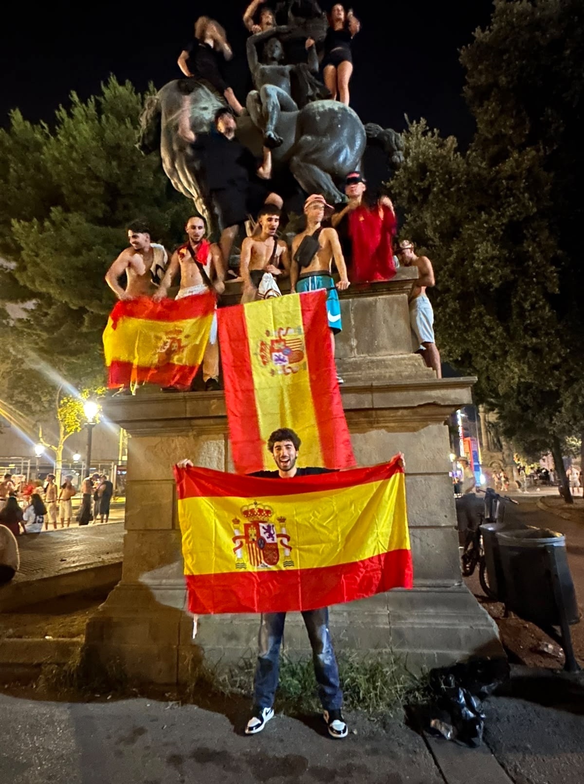 A group of men are celebrating on and around a statue at night, holding Spanish flags. One man in the foreground holds a large Spanish flag, while others are shirtless and draped in flags.
