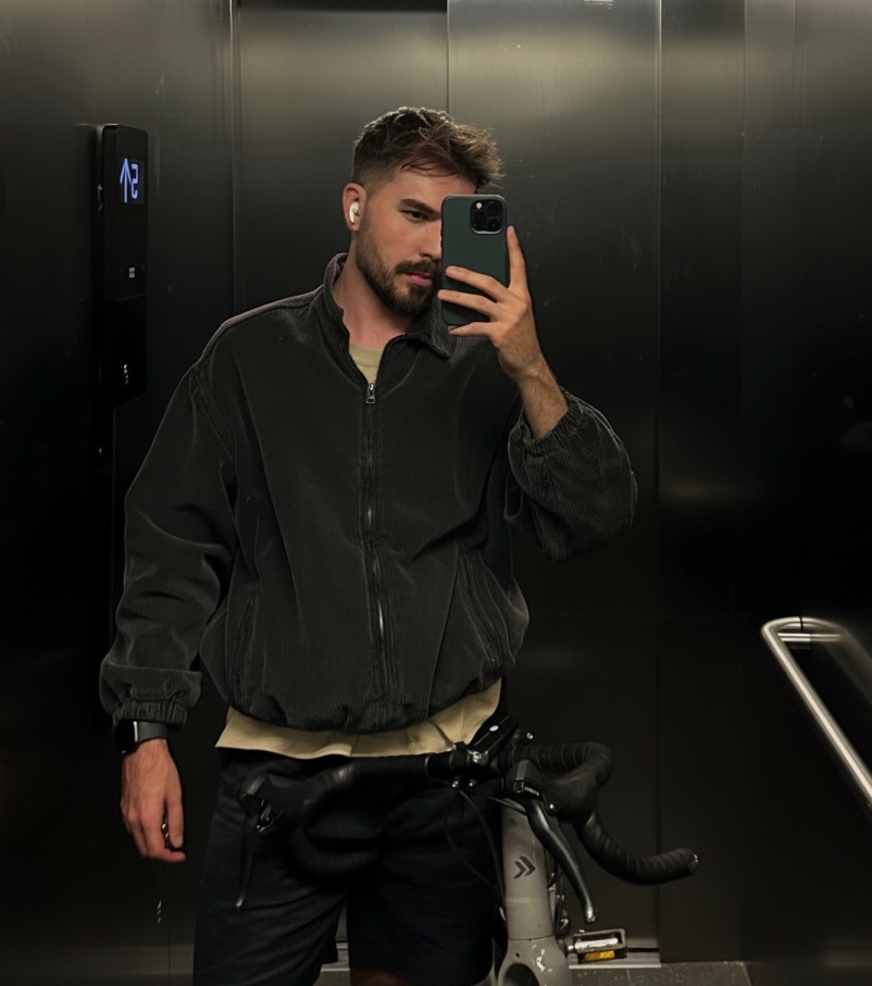 A man takes a mirror selfie in an elevator, holding his phone. He is wearing a jacket and has a bike in front of him.