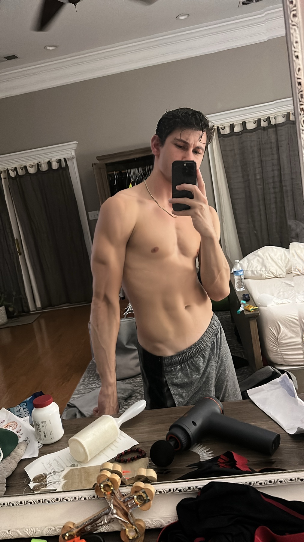 A man takes a mirror selfie in a bedroom, showing off his muscular physique. He is shirtless and wearing athletic shorts, with a massage gun and other items visible on the dresser.