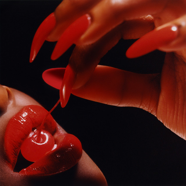 A close-up shot shows a hand with long, red, pointed nails reaching towards a mouth with glossy red lips. The hand is holding a cherry with a stem, which is dangling over the lips.