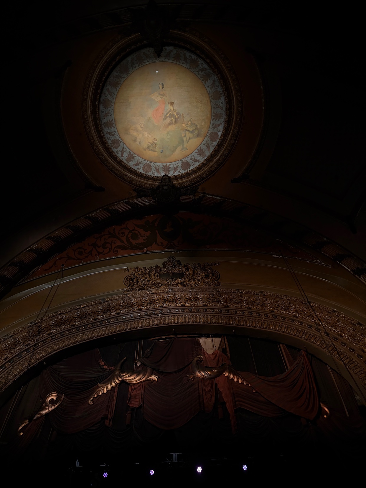 A low-angle shot of a theater's interior, showcasing its ornate ceiling and stage. The image captures the architectural details and artistic elements of the space.