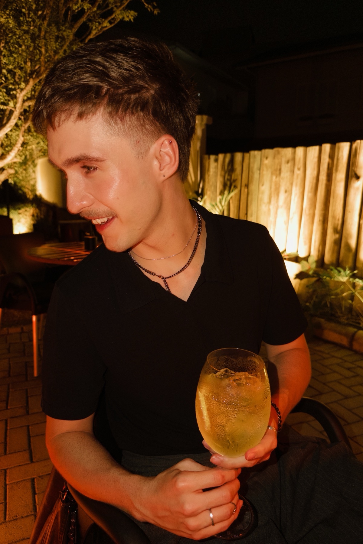 A man in his thirties smiles as he holds a large glass of a golden-hued cocktail. He is wearing a black collared shirt and appears to be at an outdoor venue at night.