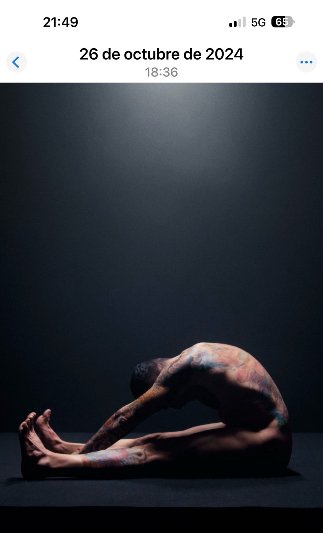 A man with body art stretches on a dark background, illuminated by a spotlight from above. His body is contorted in a seated position, showcasing his flexibility and the artistic paint on his skin.
