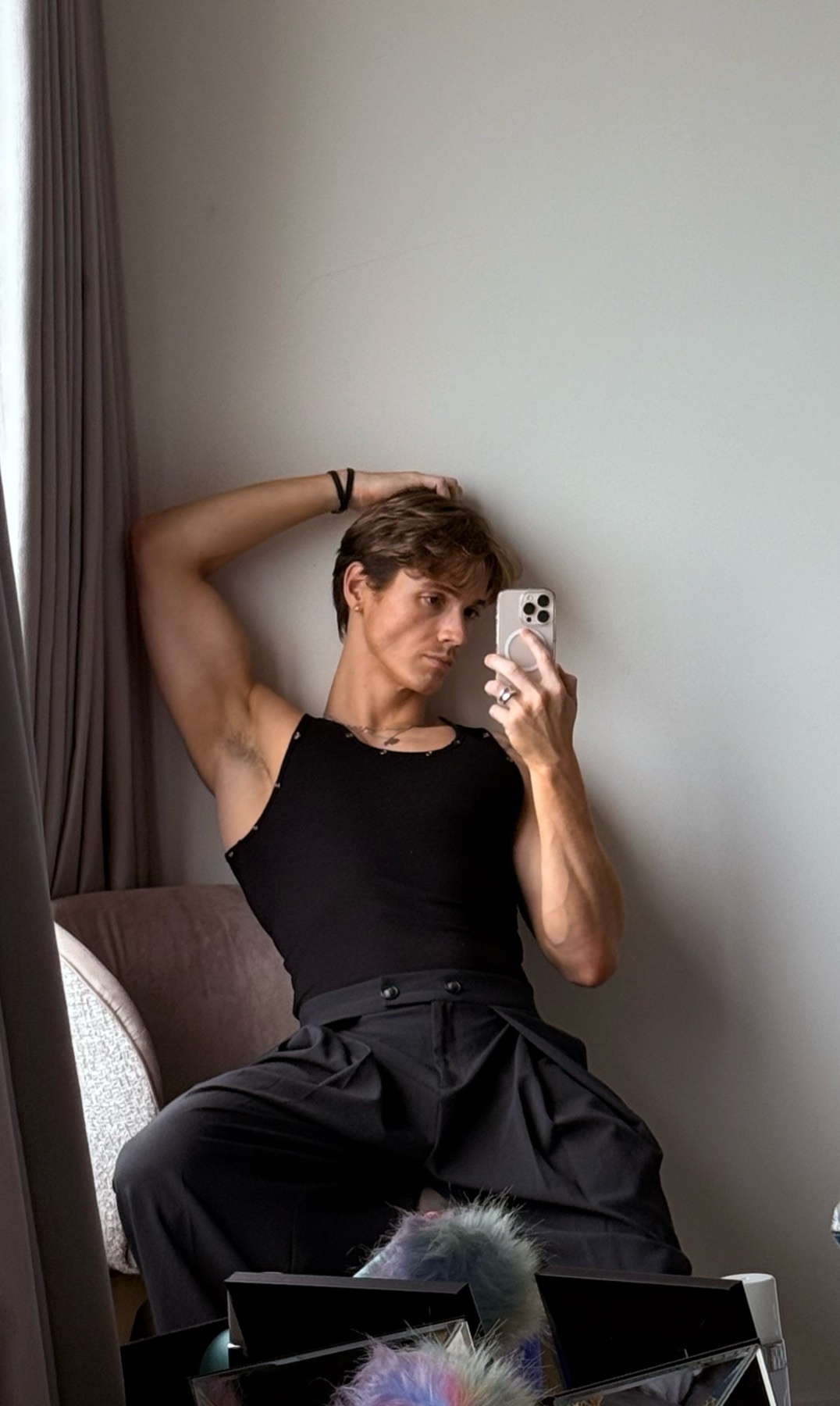 A man in a black tank top and stylish black pants poses for a selfie, holding his phone up to take the picture. He is leaning against a chair with one arm behind his head.