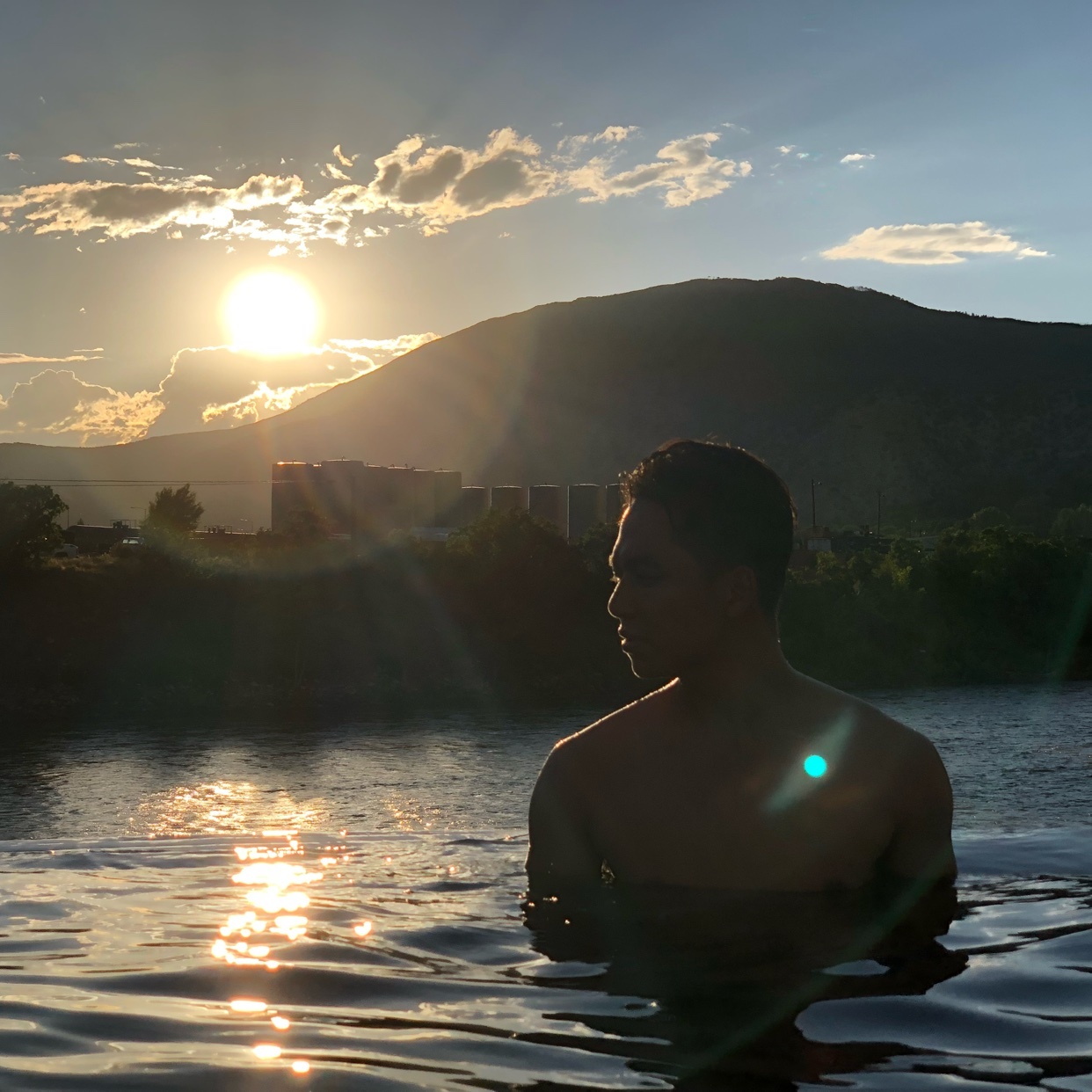 A shirtless man is in the water, looking towards a mountain range as the sun sets behind it. The sun's reflection shimmers on the water's surface.