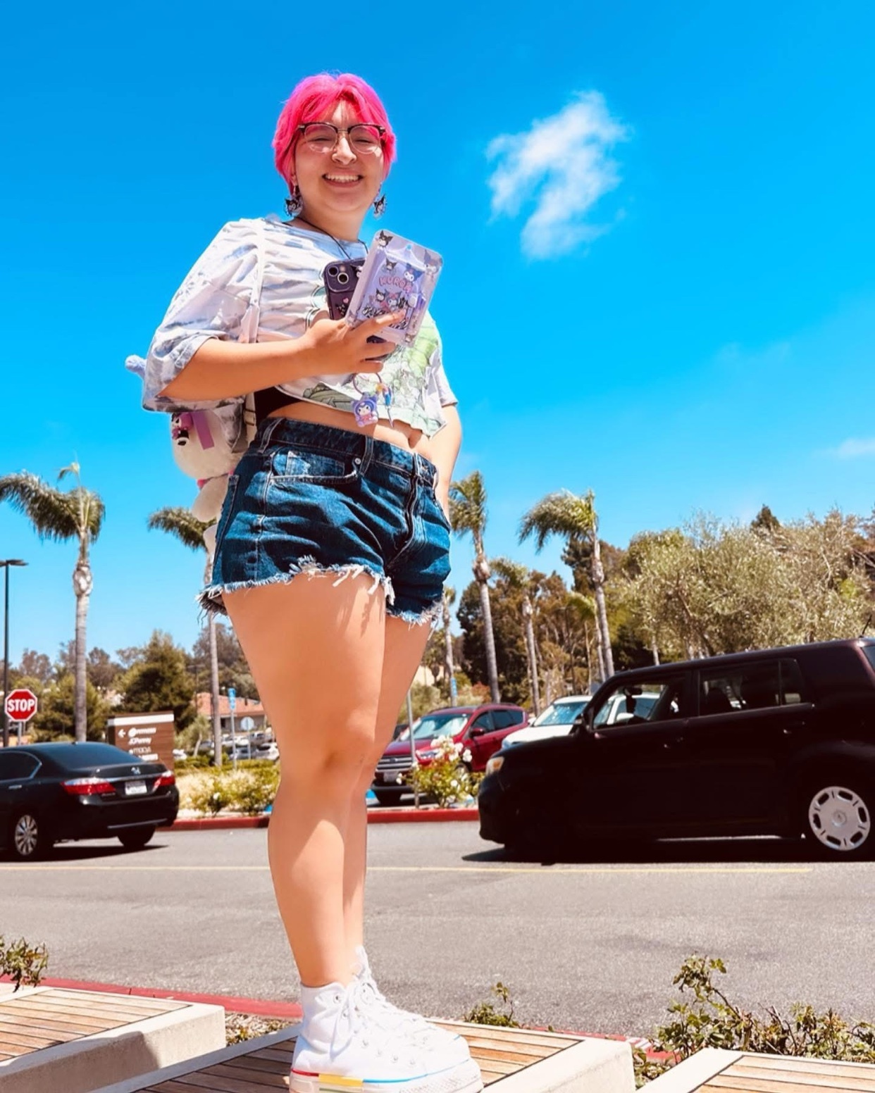 A young woman with bright pink hair and glasses poses outdoors on a sunny day. She is wearing denim shorts and a patterned top, holding a phone in her hand.