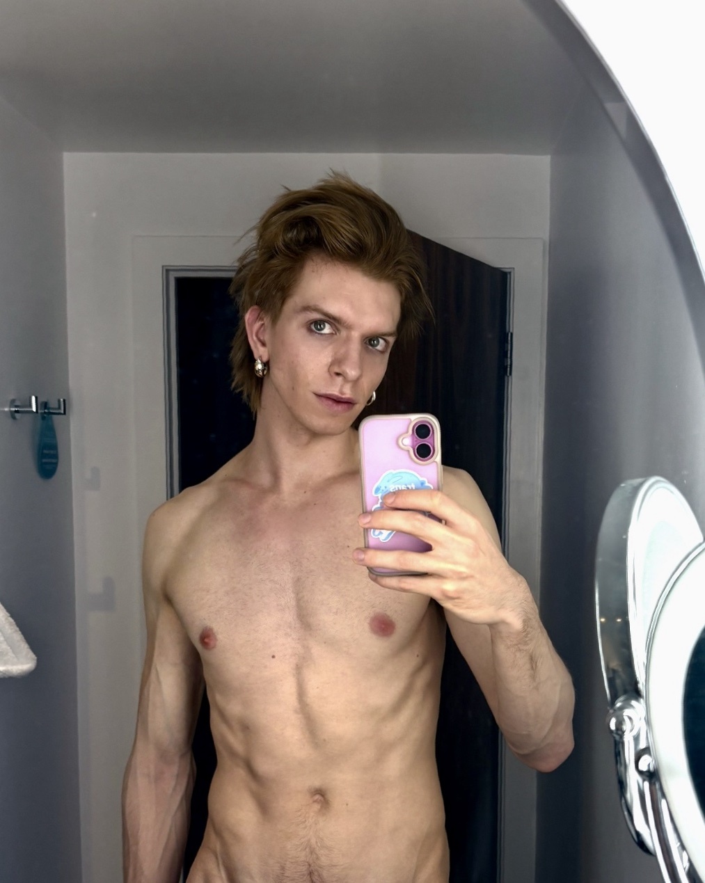 A shirtless man with tan skin and tousled brown hair takes a mirror selfie in a bathroom. He is wearing an earring and has a piercing in his nose.