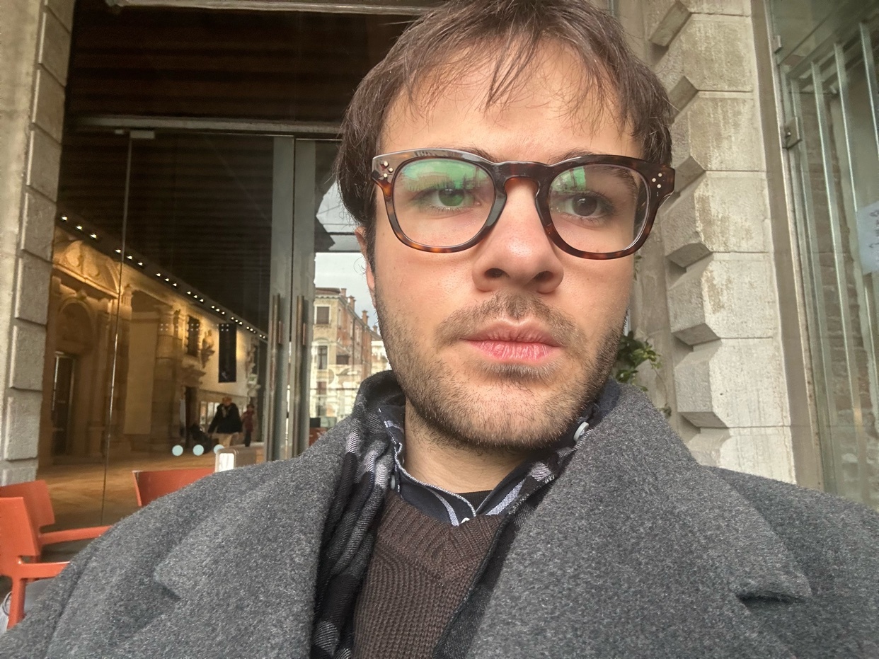 A man takes a selfie in Venice, Italy. He is wearing glasses and a coat.