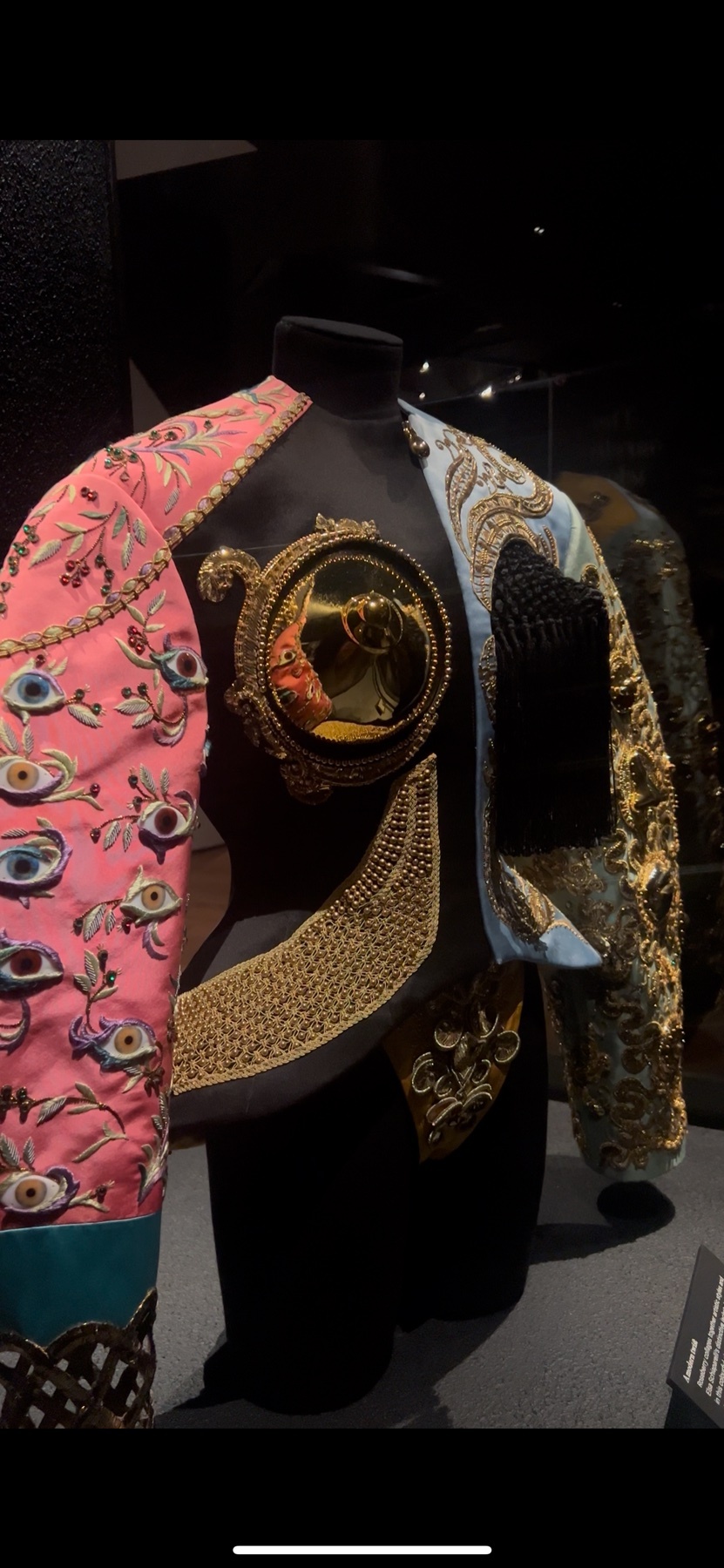 A pink and blue jacket with intricate gold embroidery and an eye motif is displayed on a mannequin in a museum setting. The ornate piece showcases Schiaparelli's signature surrealist style.