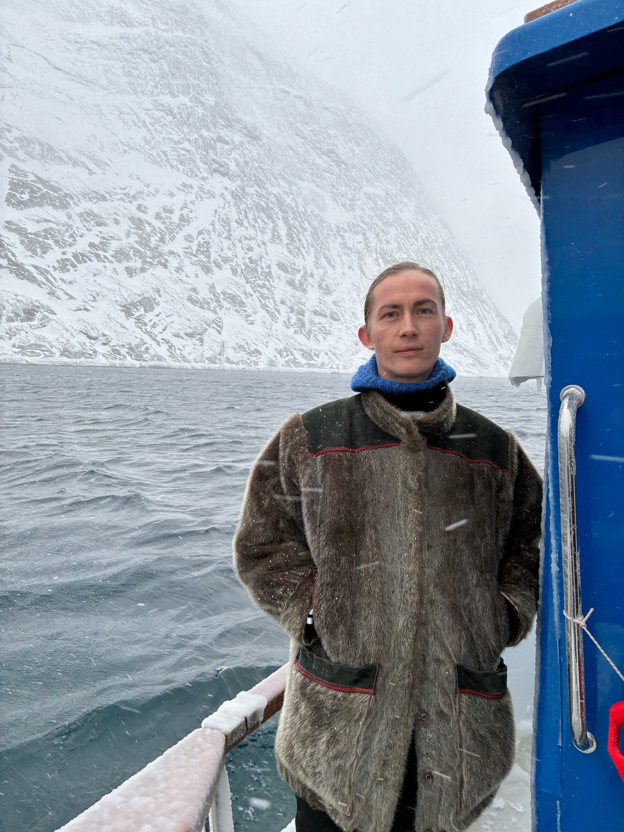 A man stands on a boat in a snowy landscape. He is wearing a fur coat and looking at the camera.