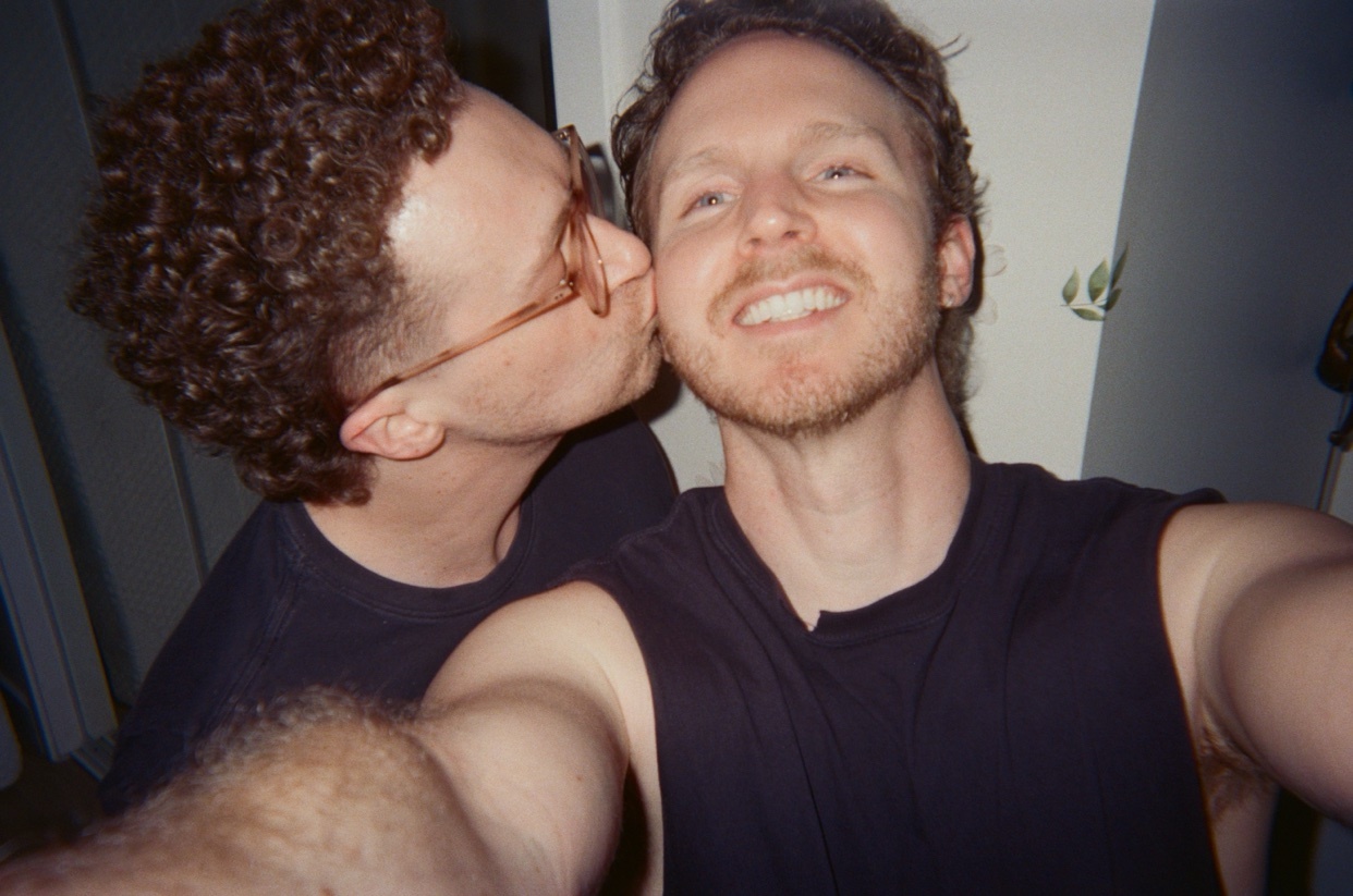 A man with curly hair and glasses kisses another man on the cheek in a selfie.