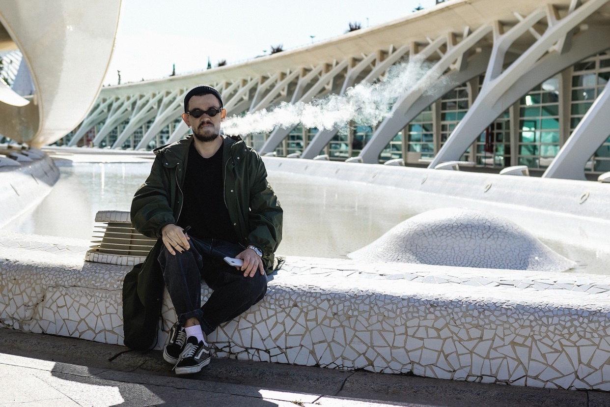 A man sits outdoors in a modern architectural setting, exhaling a plume of smoke. He wears sunglasses and a stylish green jacket over a black shirt, with dark pants and sneakers.