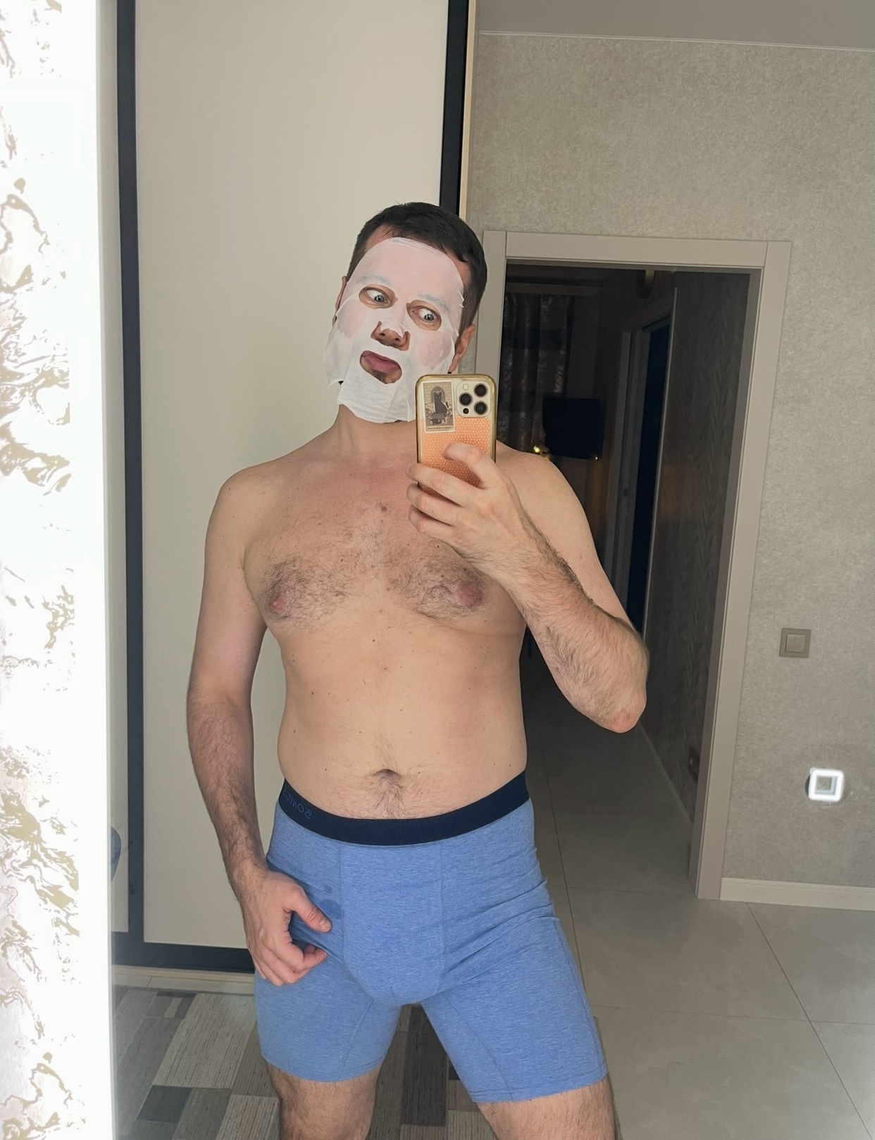 A man takes a selfie while wearing a facial mask and blue underwear. The man is shirtless and making a funny face.