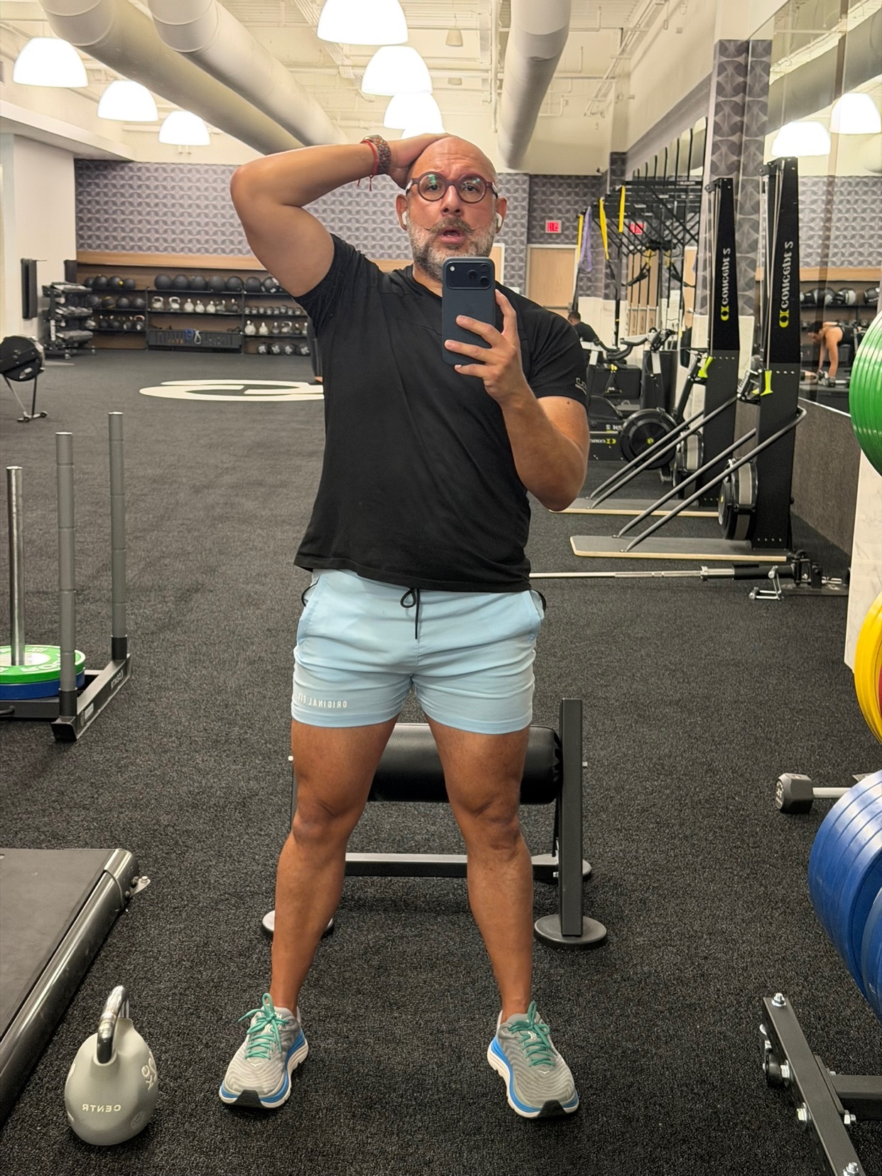 A man with glasses and a bald head poses for a selfie in a gym. He is wearing a black t-shirt and light blue shorts, with athletic shoes on his feet. The gym equipment is visible in the background.