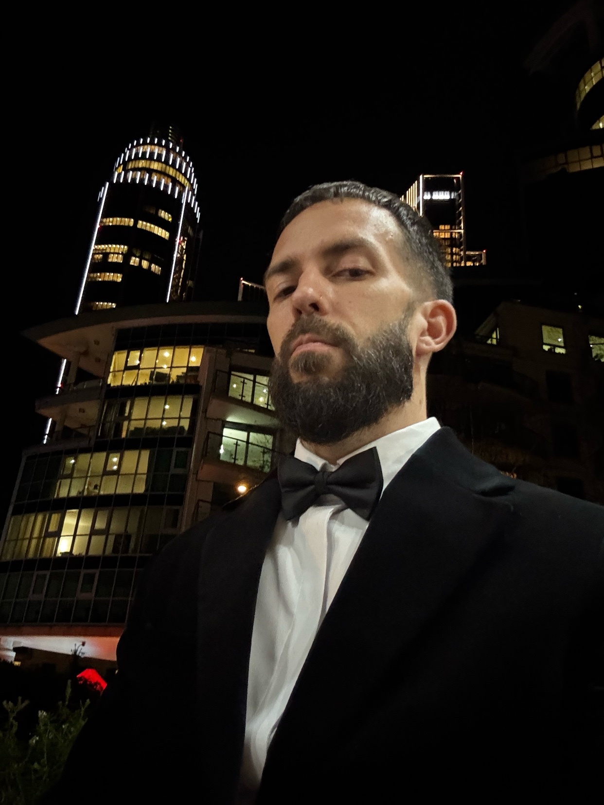 A man in a suit and bow tie poses for a photo at night. The background features illuminated skyscrapers.