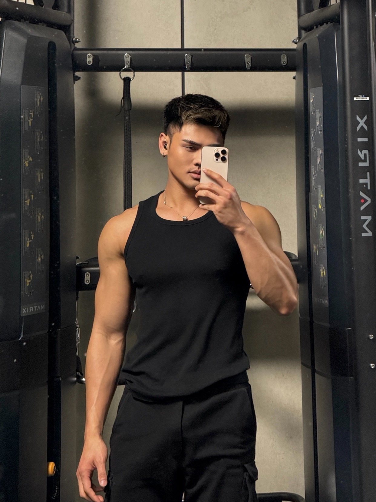A man with dark hair and a muscular physique takes a selfie in a gym. He is wearing a black tank top and black pants, posing in front of gym equipment.