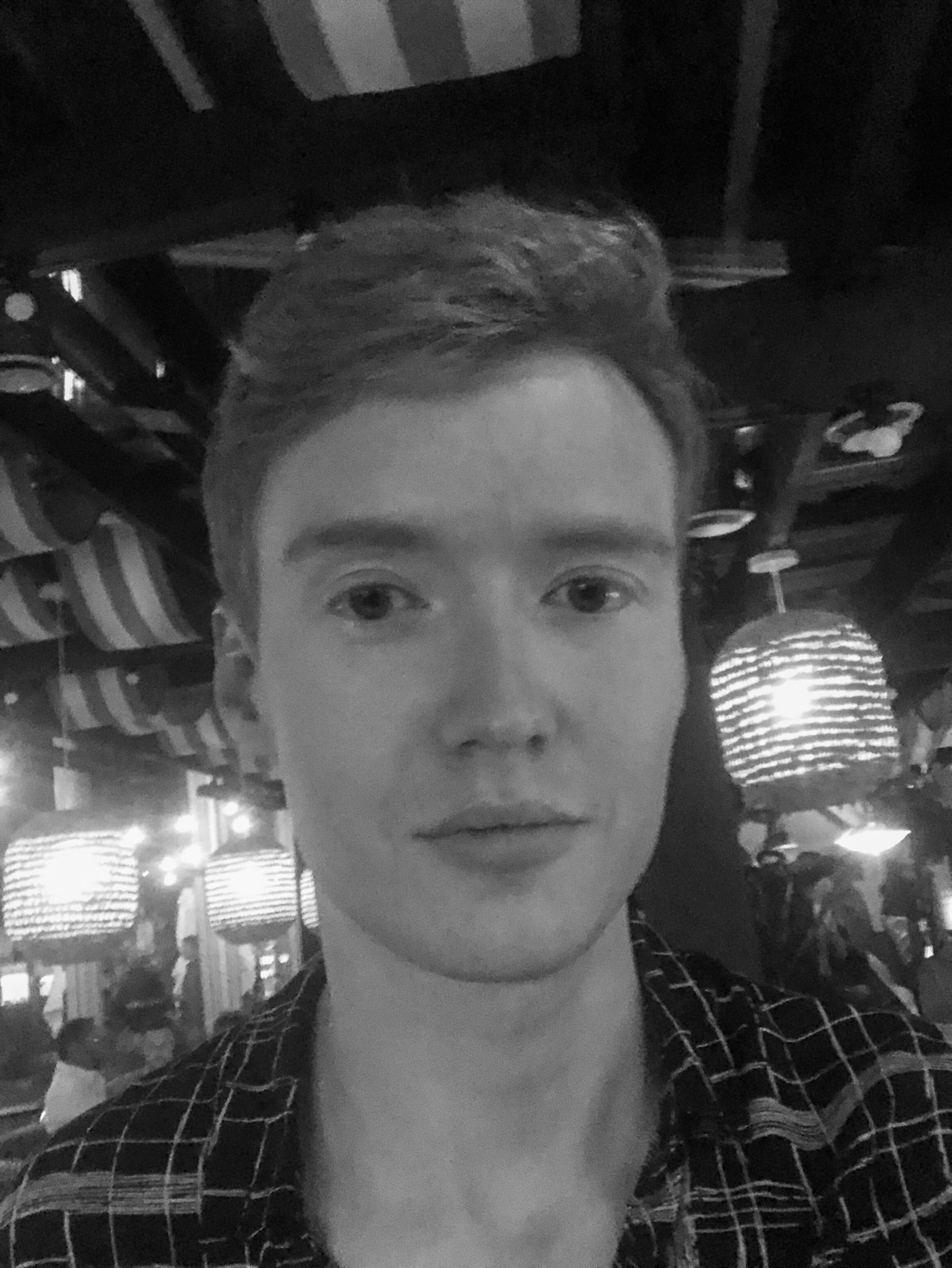 A black and white selfie of a young man with short, styled hair. He is wearing a plaid shirt and looking directly at the camera.