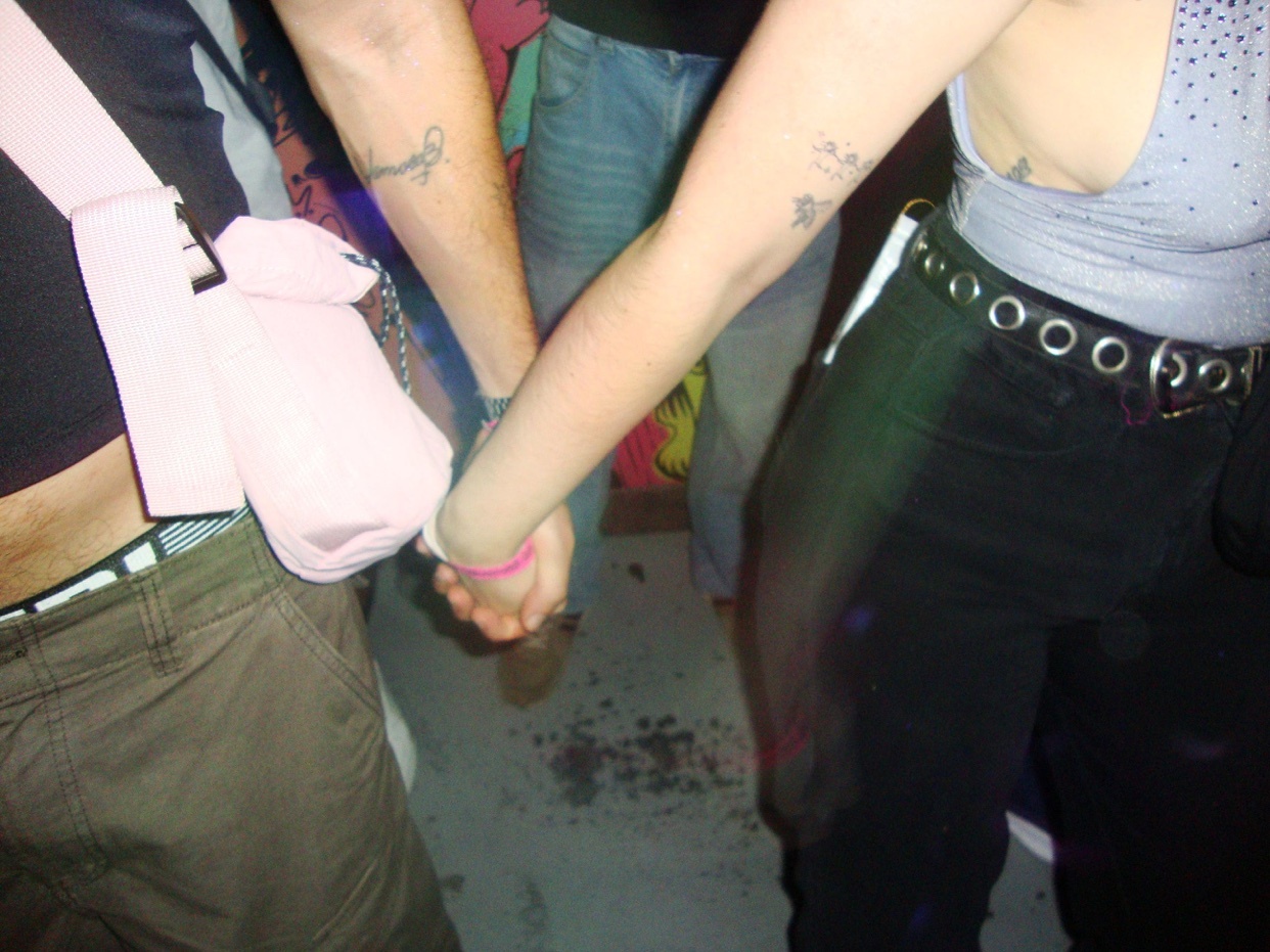 A close-up shot shows two people holding hands, with a third person visible in the background. The focus is on their arms and hands, with tattoos visible on one of the arms.