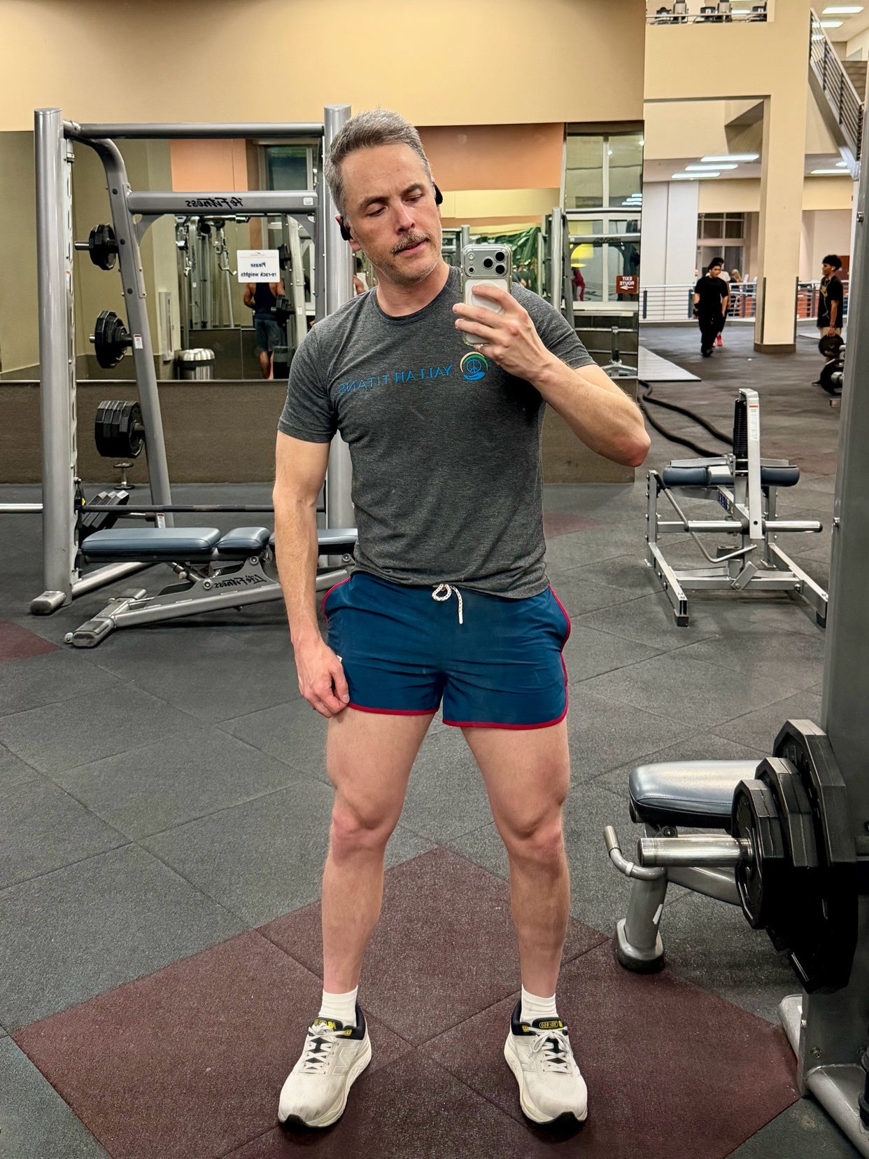 A man takes a selfie in a gym, wearing a t-shirt and shorts. He has earbuds in and a mustache.