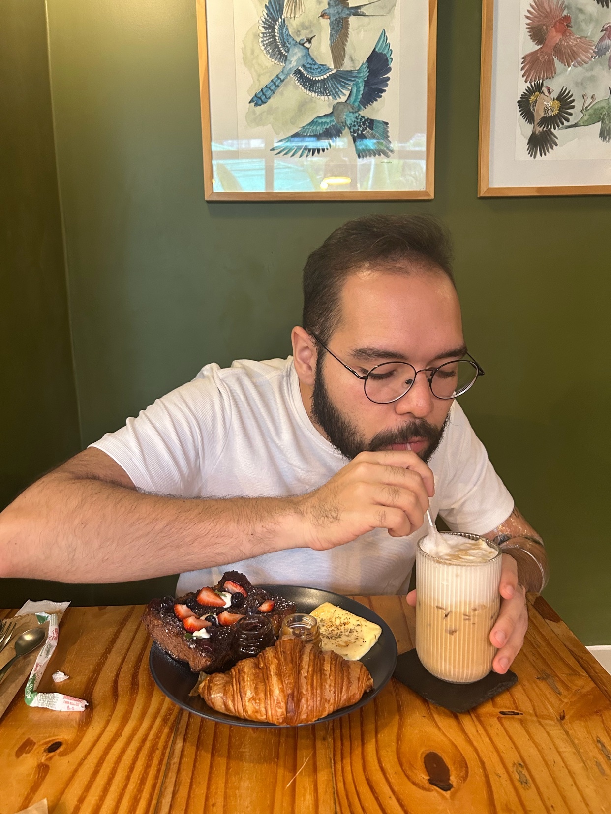 A man with glasses and a beard sips an iced coffee while sitting at a table with a plate of French toast, a croissant, and a small side of eggs. The man is wearing a white t-shirt and is seated in front of two framed pieces of art on a green wall.