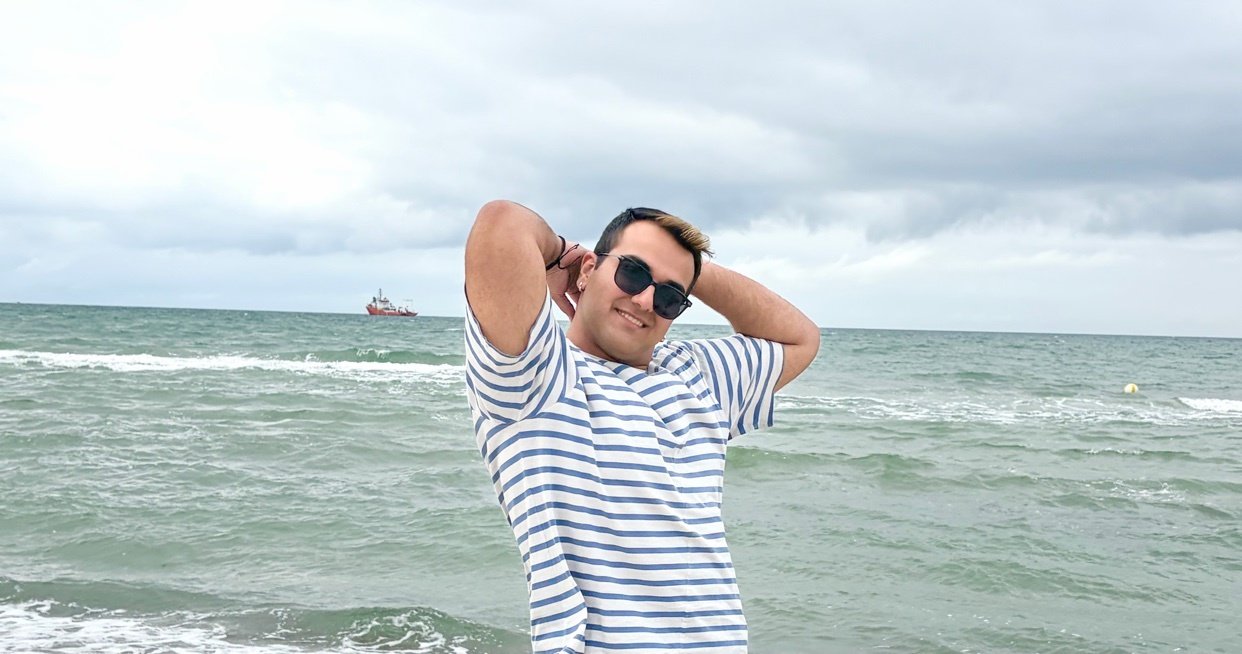 A man wearing sunglasses and a striped t-shirt poses with his hands behind his head at the beach with the ocean in the background.
