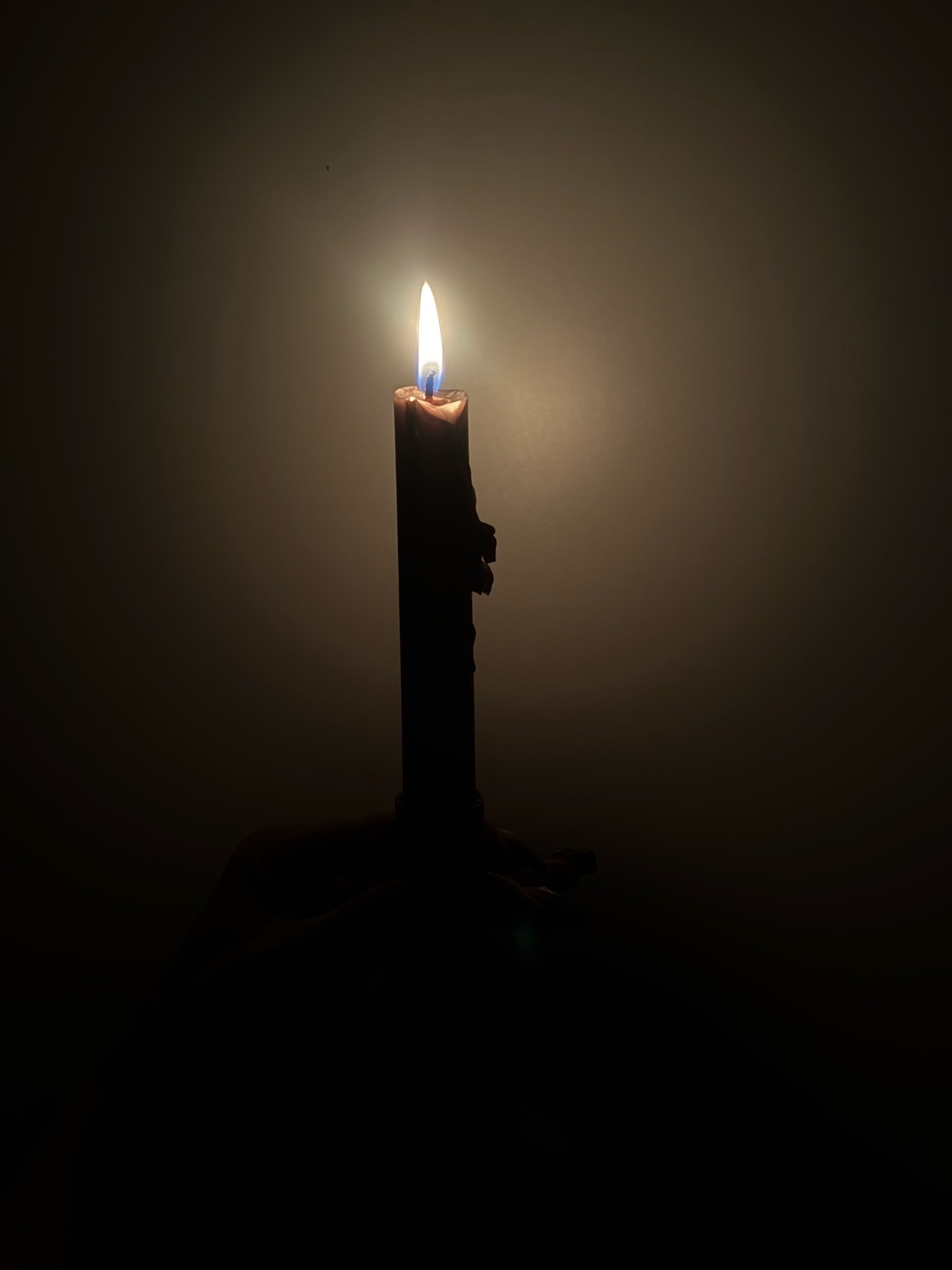 A single lit candle stands in the darkness, its flame casting a warm glow against a muted background. The candle appears to be dripping wax, adding a rustic texture to the scene.